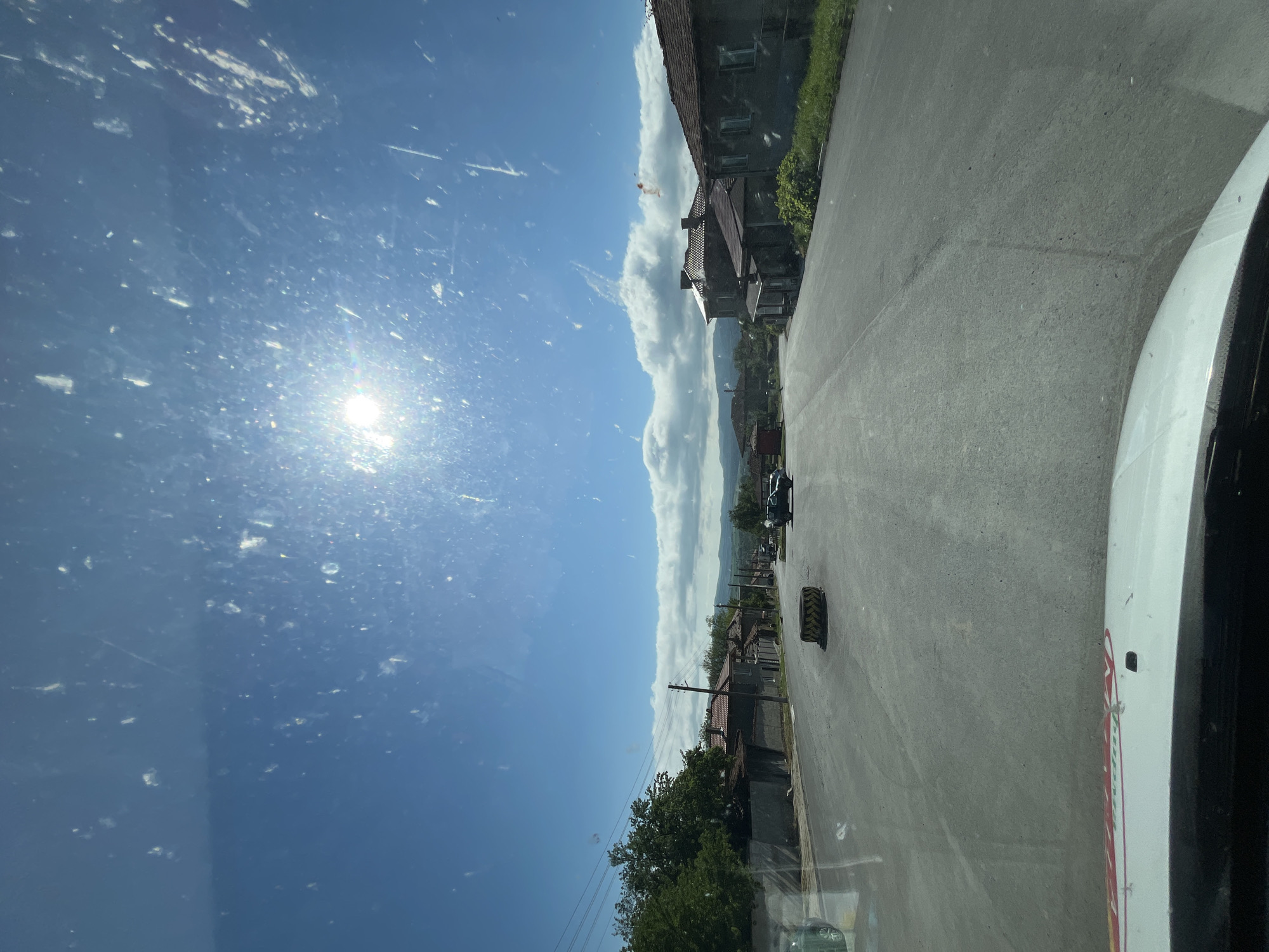 The image depicts a street scene in the city of Rish, Bulgaria, taken from inside a vehicle. The street is paved and asphalt. A discarded tire sits on the right side of the road near the foreground. Buildings line the street, varying in style and age, with trees and foliage visible in front of some of the structures. Overhead power lines run parallel to the street. A vehicle is driving along the street. The sky above is blue with clouds, and the sun glares brightly. The windshield of the vehicle appears to have dirt and streaks. The passenger-side portion of the car is visible to the right side of the image, the red and white car door on the right is of a van.