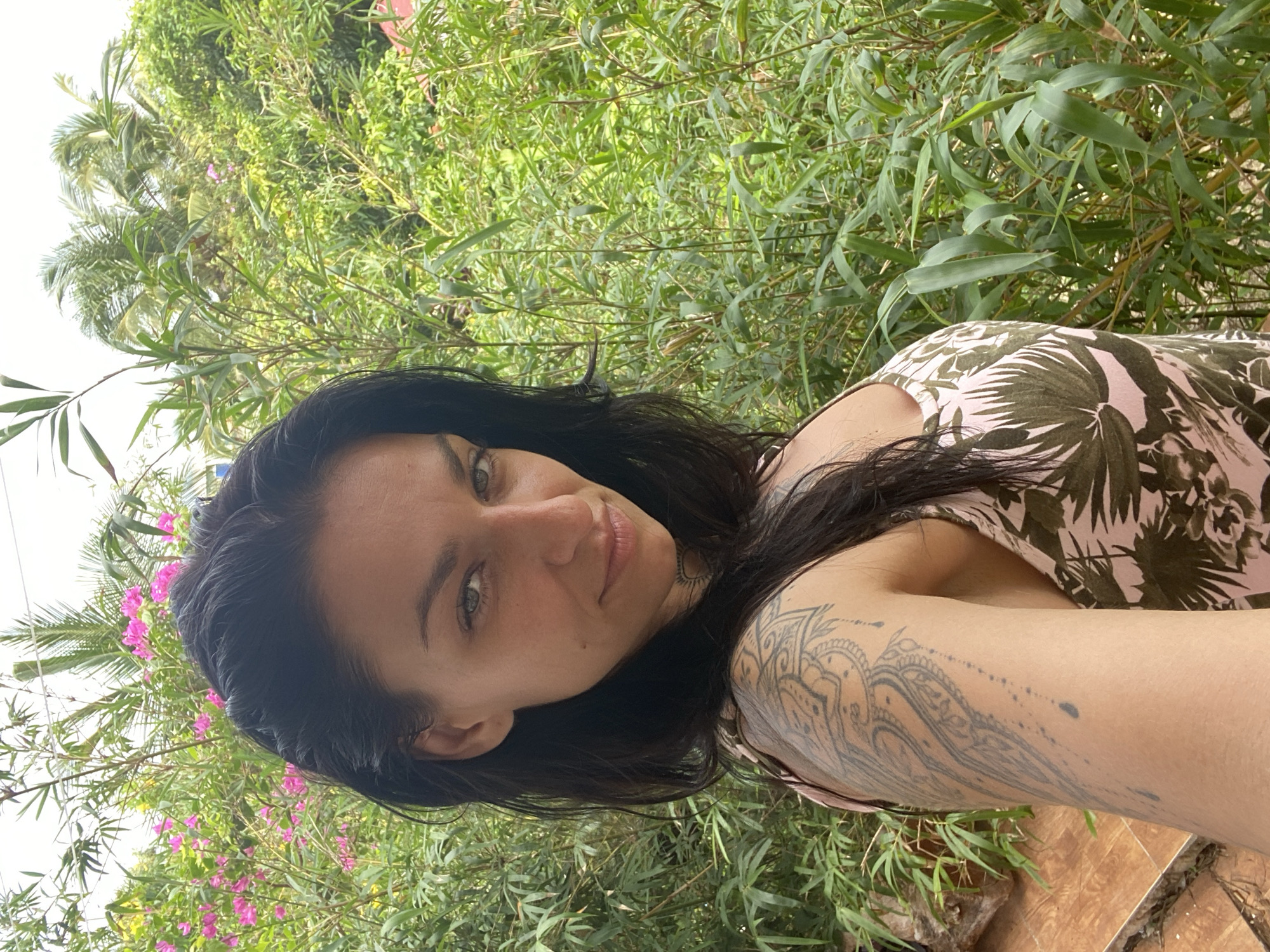A woman with dark hair and a tattoo on her left arm is taking a selfie outdoors. She is wearing a pink tank top with a green leaf pattern. The background is filled with lush green foliage, including bamboo and palm trees, with some pink flowers visible. The overall impression is of a tropical environment, consistent with a location like Mũi Né, Vietnam, known for its beaches and coastal vegetation. The lighting suggests it is daytime.