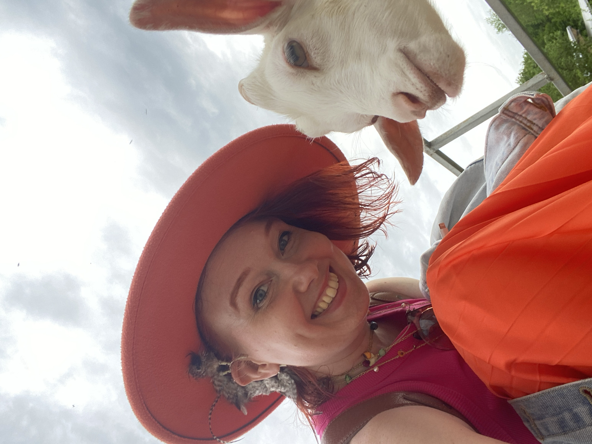 A low-angle shot captures a woman and a goat. The woman wears an orange hat and smiles at the camera. The goat is above her, seemingly peering into the frame. The background shows a cloudy sky and some trees, suggesting an outdoor setting. An orange jacket and other accessories are visible on the woman.