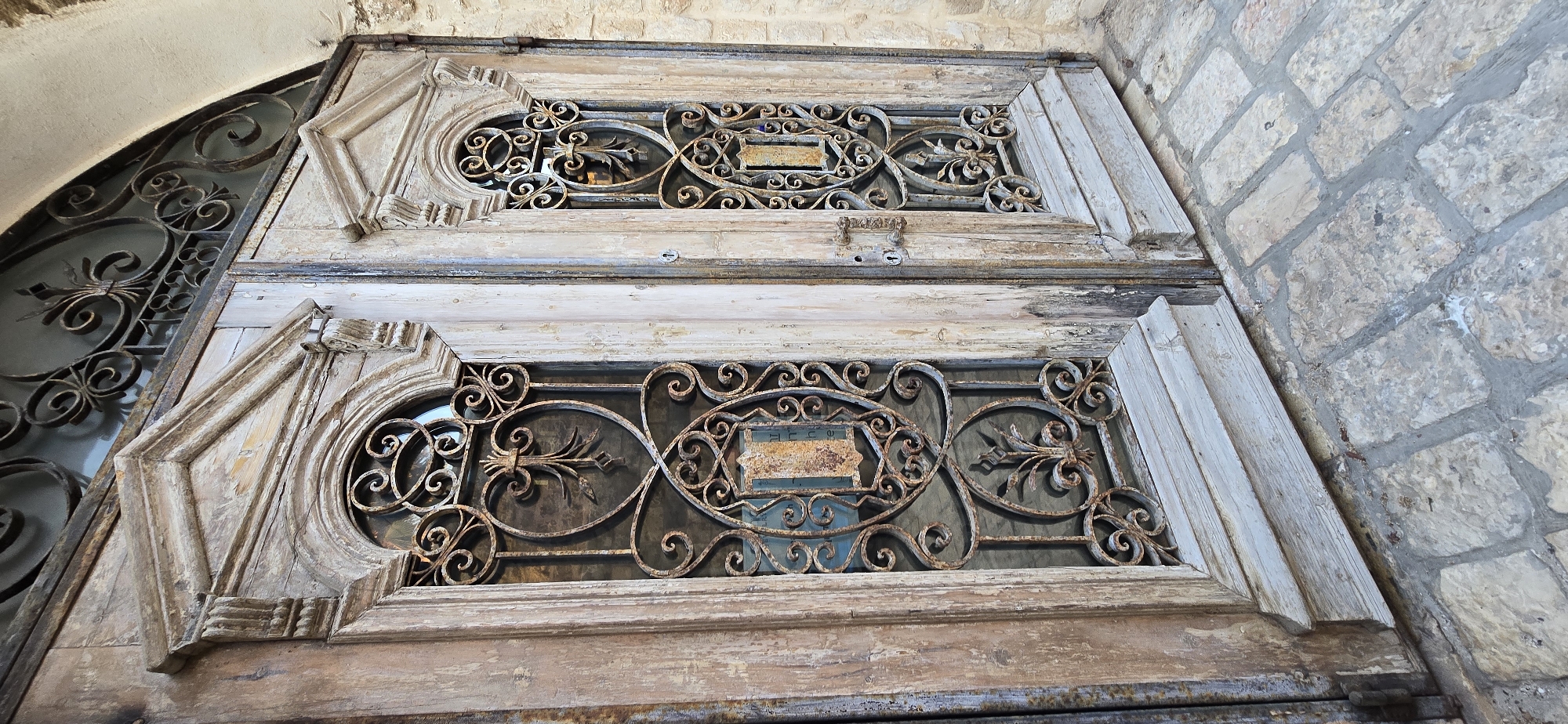The image shows a weathered, light-colored wooden double door with ornate, rust-colored wrought iron inserts. The doors are set within a stone wall and open onto a cobblestone pathway. The wrought iron features intricate scrollwork, floral motifs, and shield-like designs. The wood of the doors shows signs of age and wear, with peeling paint and visible grain. The overall scene suggests an old, possibly historic, building entrance. The location is identified as Tel-Aviv, Israel. There are no people or specific actions depicted in the image.