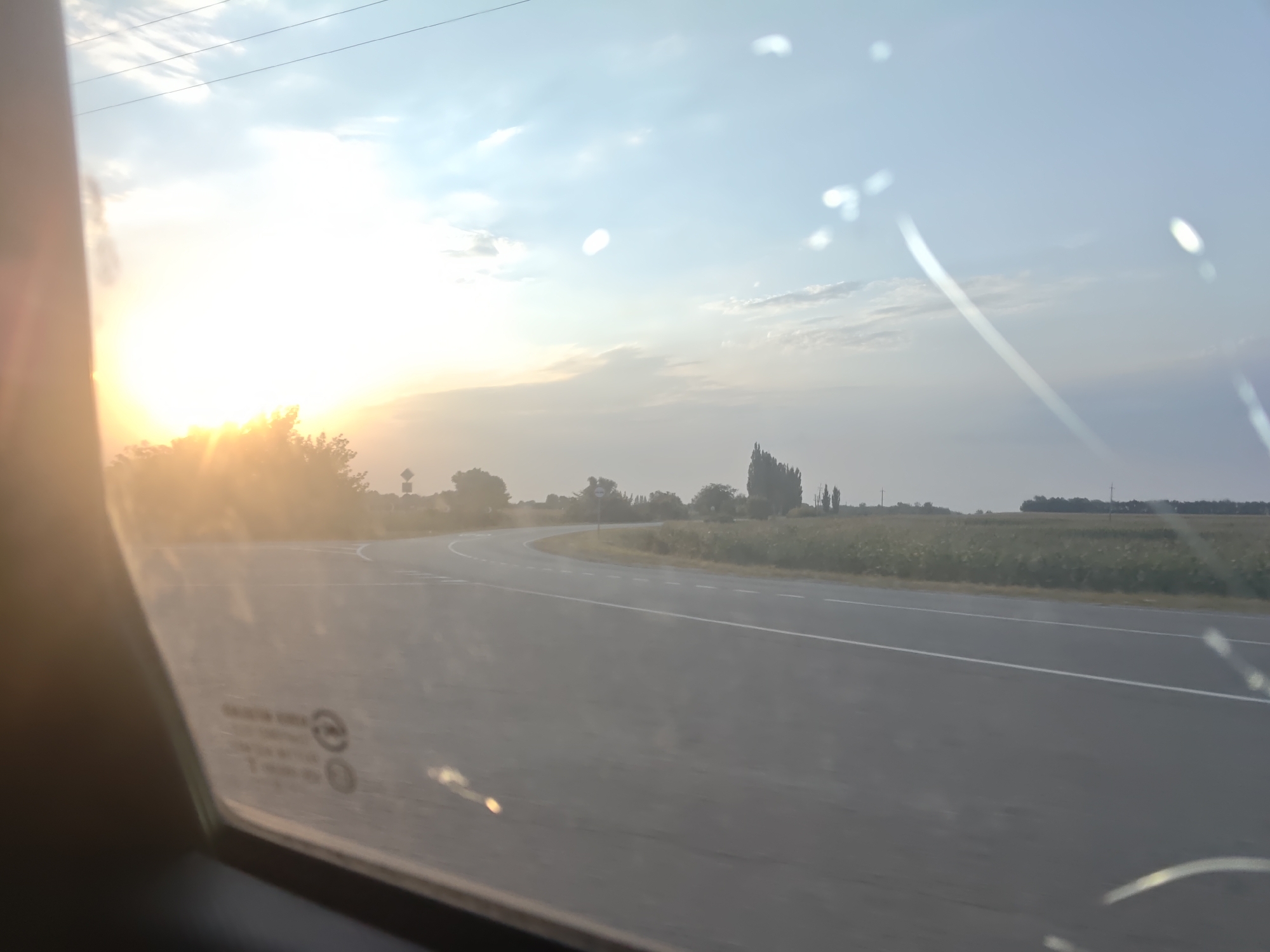 The image shows a view from inside a vehicle, looking out through a slightly dirty car window at a rural landscape. The sun is low on the horizon, casting a bright glare and lens flare across the scene, suggesting it is either sunrise or sunset. A curved asphalt road with white lane markings stretches into the distance, bordered by fields and scattered trees. The sky is partly cloudy with a pale blue hue. The window itself has several smudges and a few fine scratches. In the lower left corner of the window, a faint imprint of car window safety information and certification marks is visible. The overall impression is of a journey through a peaceful, rural environment in the early morning or late evening. There are no people or animals visible in the frame, and no specific landmarks are identifiable to pinpoint the exact location within Veselyi, Russia.