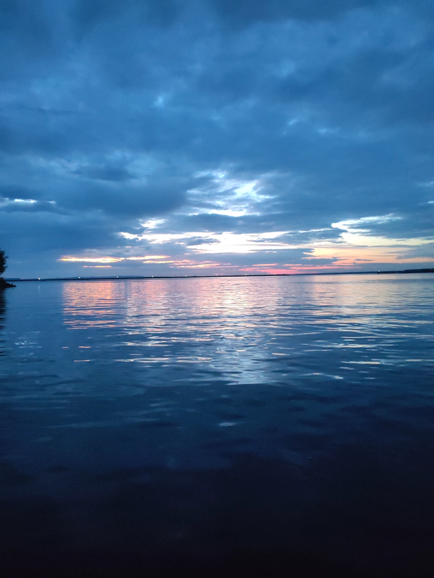 The image depicts a wide body of water, likely a river or lake, under a dramatic, cloudy sky. The clouds are a deep blue, with streaks of lighter blue and white where the light is breaking through. Towards the horizon, there is a band of soft orange and pink light, indicating either sunrise or sunset. This light is reflected in the rippling surface of the water, creating shimmering patterns of color. The water itself is a deep blue, transitioning to near black in the foreground, suggesting depth and possibly the absence of light penetration. On the far left edge of the image, a dark, silhouetted landmass with some vegetation can be seen. The scene conveys a sense of calm and natural beauty, typical of twilight hours. This view is likely of the Kama River in Chaikovsky, Russia, known for its scenic waterfronts, especially during dawn and dusk. There are no people or man-made objects clearly visible in this landscape.
