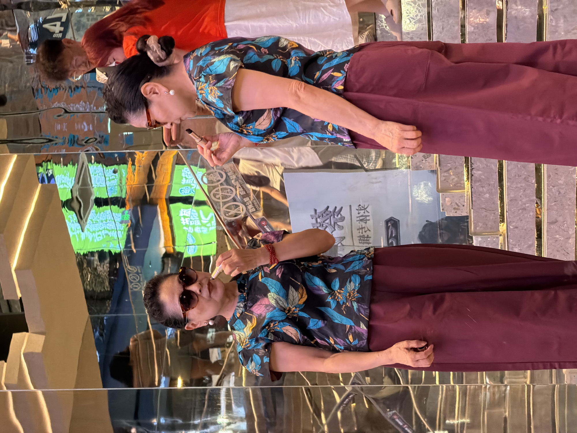 The image depicts two women applying lipstick in front of a mirrored wall with neon lights reflecting on it. The woman on the left is wearing sunglasses and a floral print top with puff sleeves and maroon pants. She has a red bracelet on her wrist. The woman on the right is wearing a similar top and maroon pants, and she has a pearl earring. Behind them, other people are visible, including a man with glasses and a woman with red hair. The setting appears to be an urban environment, possibly a shopping district in Yuzhong District, China, given the reflections of signage in the background. The overall atmosphere suggests a moment of personal grooming or preparation in a public space.