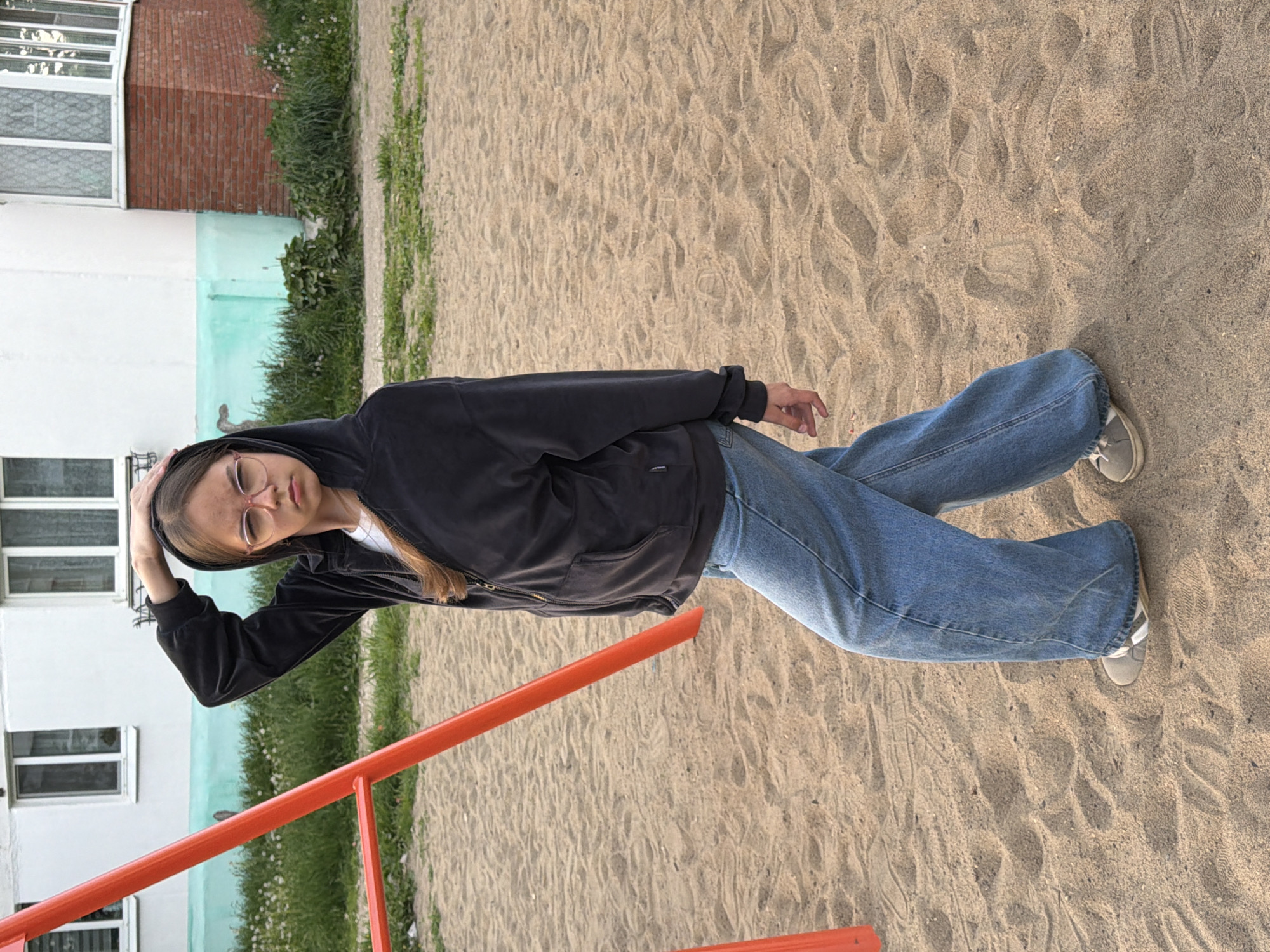 A woman is on a playground in Chaikovsky, Russia, with a sandy surface. The woman has long blonde hair, wears glasses, a black hoodie, a white shirt, and blue jeans. She is standing on a playground next to a red metal structure. A multi-story building with white window frames stands behind her, adjacent to green vegetation and lawn. The image appears to be taken from a ground-level perspective.
