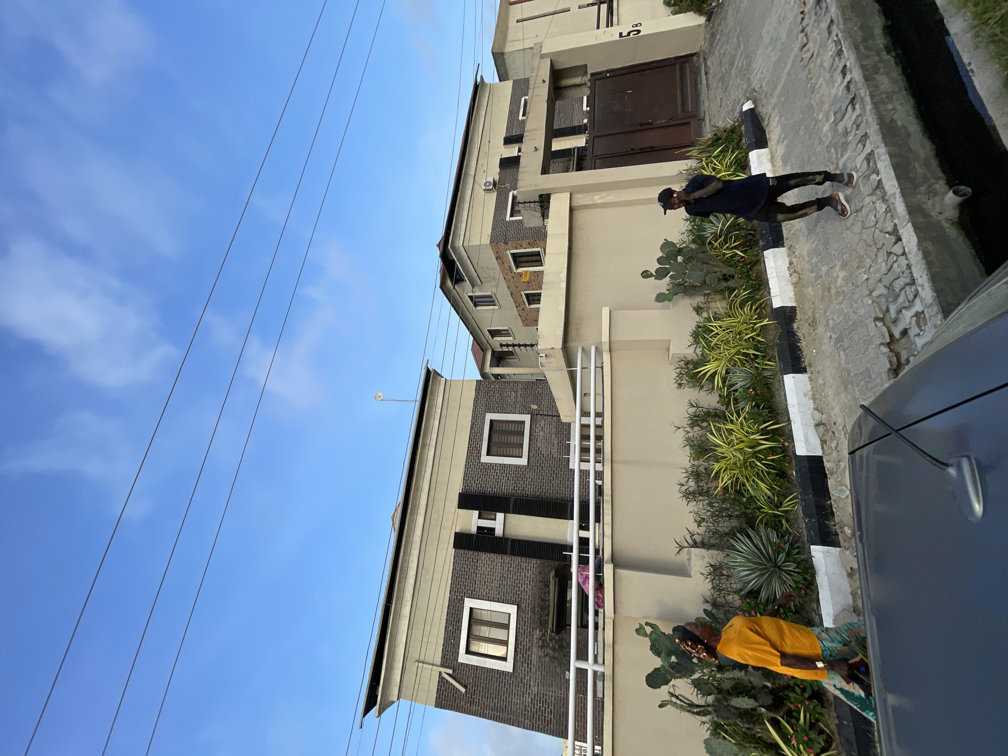 The image depicts a street scene in Ilado, Nigeria, with two individuals in the foreground and modern multi-story buildings in the background. On the left, a woman wearing a bright orange t-shirt and patterned trousers walks from left to right, carrying a dark bag. She has a headscarf with a similar pattern to her trousers. In the middle ground, a man wearing a dark blue t-shirt, patterned trousers, a black baseball cap, and flip-flops stands and appears to be speaking on a mobile phone. He is positioned on a paved walkway next to a drainage ditch.
Behind the individuals, a beige-colored wall runs horizontally across the frame, with various types of arid plants, including cacti and succulents, growing in front of it. The wall is topped with a white railing. Beyond the wall, two large residential buildings are visible. The building on the left has a dark brick facade with lighter colored horizontal banding and a slanted roofline. The building on the right is predominantly light beige with brick accents. Both buildings have multiple windows and air conditioning units. Several overhead power lines traverse the blue sky, which has scattered white clouds. A dark-colored car is partially visible in the lower foreground, obscuring the bottom edge of the frame. The overall impression is of a residential area in an urban environment.