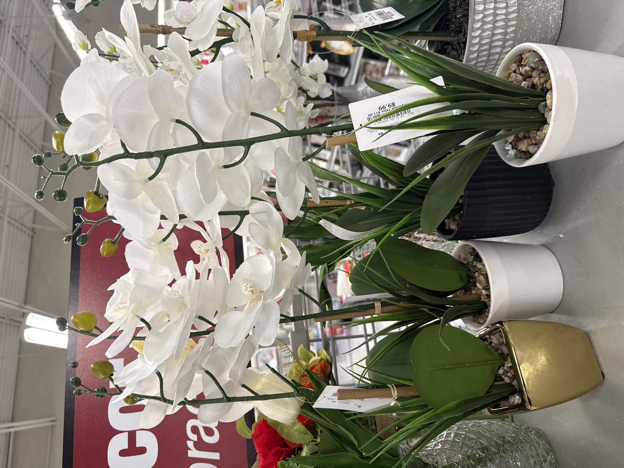 The image depicts a variety of artificial plants, including white orchids and greenery in decorative pots, arranged on what appears to be a shelf or display table. The setting likely is a retail store, possibly a home décor or craft supply shop, based on the nature of the merchandise. One of the price tags visible indicates that the plants are sold for $9.99. The photo was likely taken in Columbus, United States of America.