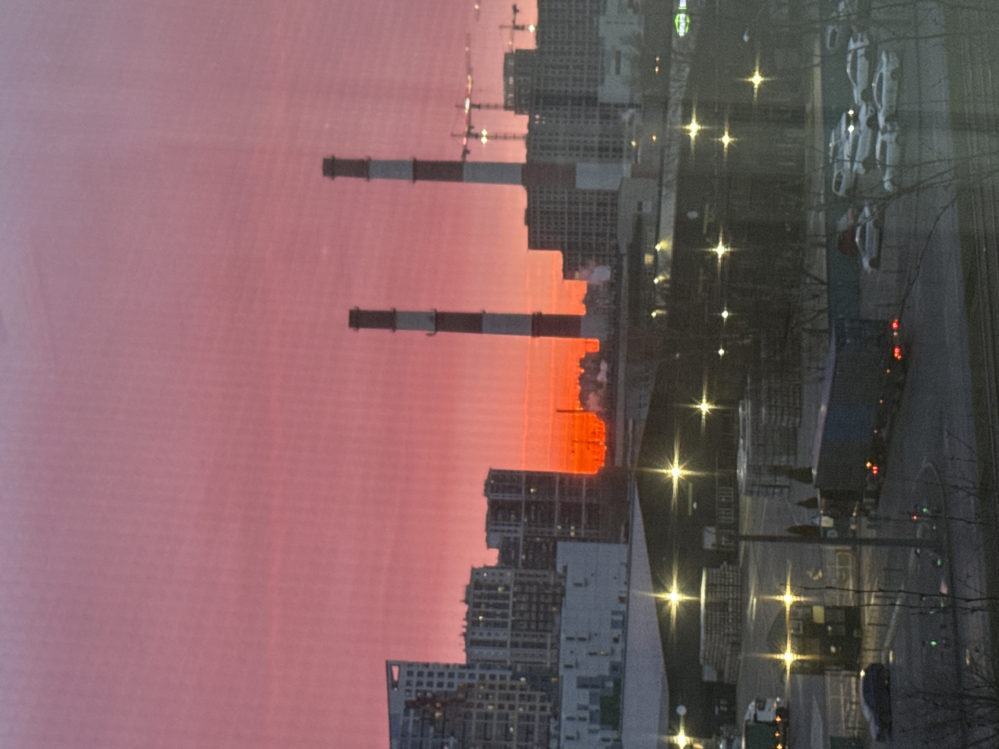 The image shows a view in Saint Petersburg, Russia, at dusk or dawn. The sky is a gradient of pink and orange. In the middle distance are industrial chimneys with black and white stripes, and nearby are modern buildings and construction cranes. On the right are streetlights, parked cars, trees, and what seems to be a parking lot.