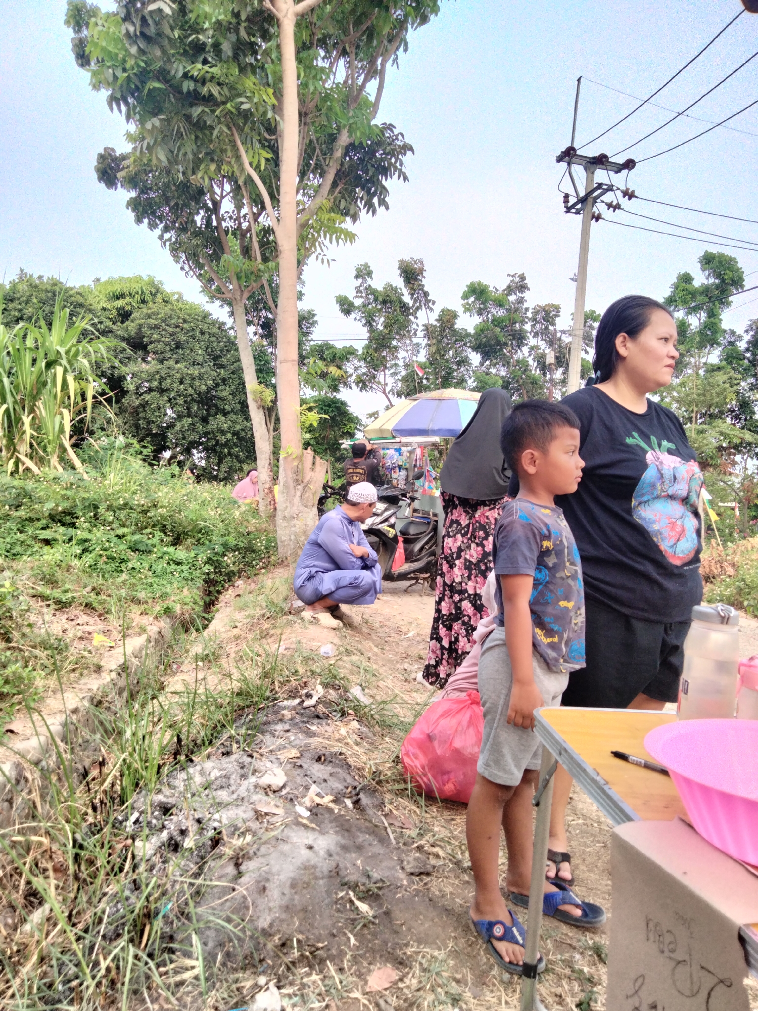 In Cariu, Indonesia, a modest outdoor scene unfolds under a clear sky, with a prominent tree on the left providing partial shade. A woman, wearing a black t-shirt with a colorful heart graphic and black shorts, stands with a boy in a grey t-shirt and grey shorts. The boy wears blue flip-flops with a Captain America emblem. They are positioned near a portable table, which holds a pink bowl, a bottle, and a pen. Further in the background, another woman in a dark headscarf and a floral dress is partially visible, standing near a small market stall with various items displayed. To the left, a man in a light blue traditional outfit and a white skullcap is crouched down, facing away from the camera. The ground is dirt and overgrown with grass and some debris. A red plastic bag is visible near the stall. Electrical poles and wires are present in the background, indicating the presence of infrastructure. The overall setting appears to be a roadside or market area in a rural or semi-urban environment.