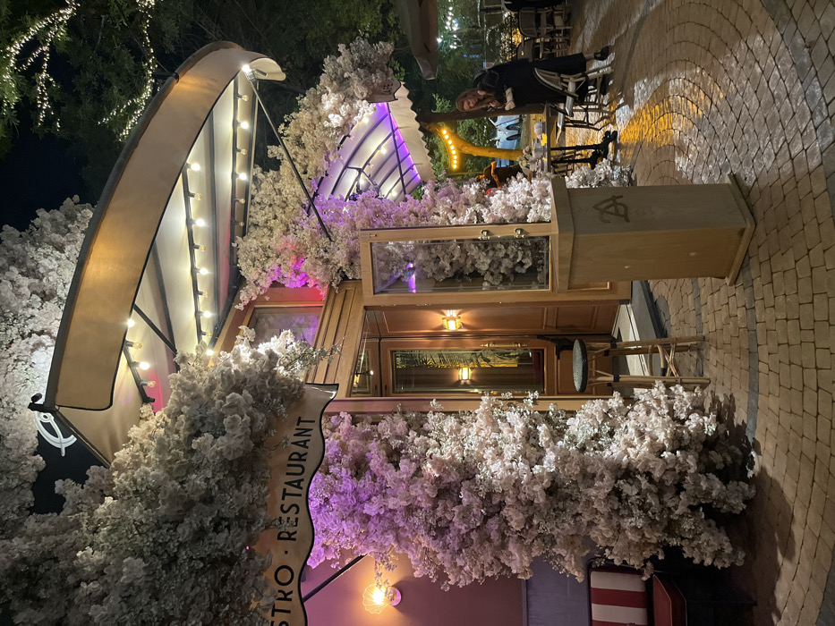 A quaint bistro restaurant, lavishly adorned with white faux flowers, occupies a corner space. A curved awning with light bulbs is partially visible. Clear glass panels filled with blossoms line the upper exterior, while a wooden counter with bar stools sits underneath. The building signage reads "Bistro Restaurant." A person stands outside, and the scene is set against a night sky with visible lighting.