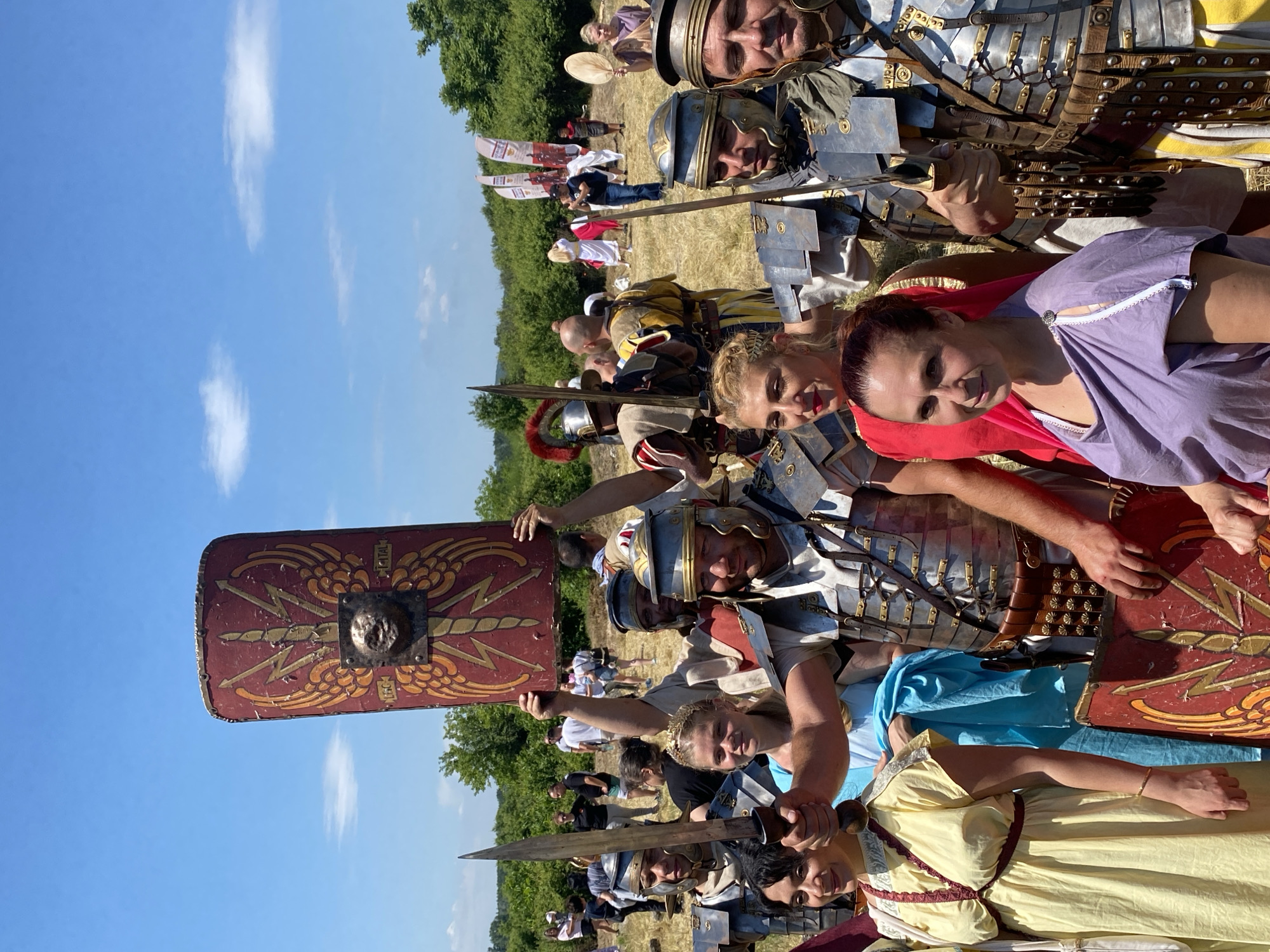 The image depicts an outdoor historical reenactment event in Gigen, Bulgaria, under a clear blue sky with scattered white clouds. In the foreground, a group of approximately ten individuals is posing for the camera. Several men are dressed in detailed Roman legionary armor, including lorica segmentata and helmets, and hold gladii (Roman short swords). One man, centrally positioned, holds a large red and gold scutum (rectangular shield) over his head, featuring winged lightning bolts, a central Gorgon or lion-like boss, and inscriptions "LEG" and "ITAL". Alongside them are several women dressed in Roman-style tunics and stolas of various colors, including yellow, red, purple, and light blue. One young woman wears a golden wreath on her head. Most individuals in the foreground exhibit pleasant expressions, with several smiling directly at the viewer. The ground consists of dry, golden grass, indicative of a field. In the midground and background, numerous other people are visible, some in casual modern clothing, others possibly in historical costumes, suggesting a public gathering or festival. A line of dense green trees and bushes forms the horizon in the distance. Two white banner flags with graphics are partially visible to the right side of the frame in the background.