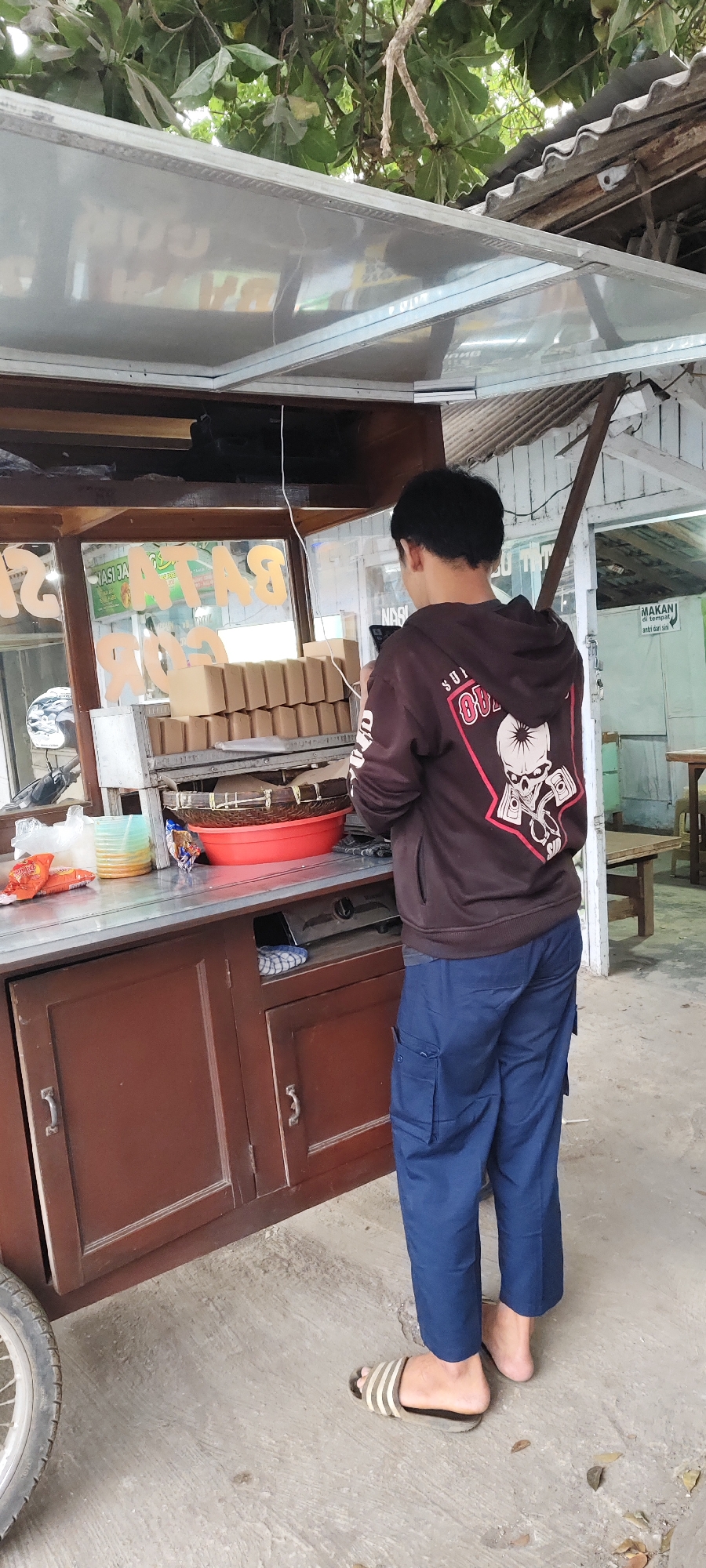 A young man, seen from behind, is standing in front of a food stall in Malang, Indonesia. He is wearing a brown hoodie with a graphic design of a skull and pistons on the back, and blue cargo pants. He is holding a smartphone in his right hand and appears to be looking at it. The food stall is made of wood and metal, with a transparent roof. It is stocked with boxes of food, a red bowl, and various other items. In the background, there is a building with a sign that says "MAKAN", which means "eat" in Indonesian. There are also tables and chairs outside the stall. The ground is paved with concrete, and there are some leaves scattered around. The overall scene suggests a casual outdoor dining setting.