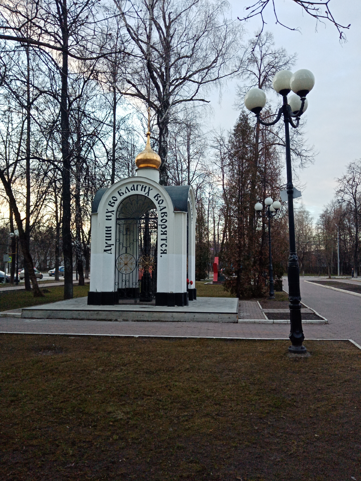 The image shows a small, white-walled chapel with a golden dome, located in a park in Elektrostal, Russia. The chapel has arched entrances with black metal gates adorned with circular patterns. Cyrillic text is painted vertically on the sides of the chapel. A black curb runs along the base of the structure. The chapel is built on a concrete platform that extends into a paved area.
The surrounding environment includes bare trees, lawns with brown grass, a lamppost with four globe-shaped lights, and paved walkways. Other trees and a monument are visible in the background, as are cars in the distance. The sky is overcast.
