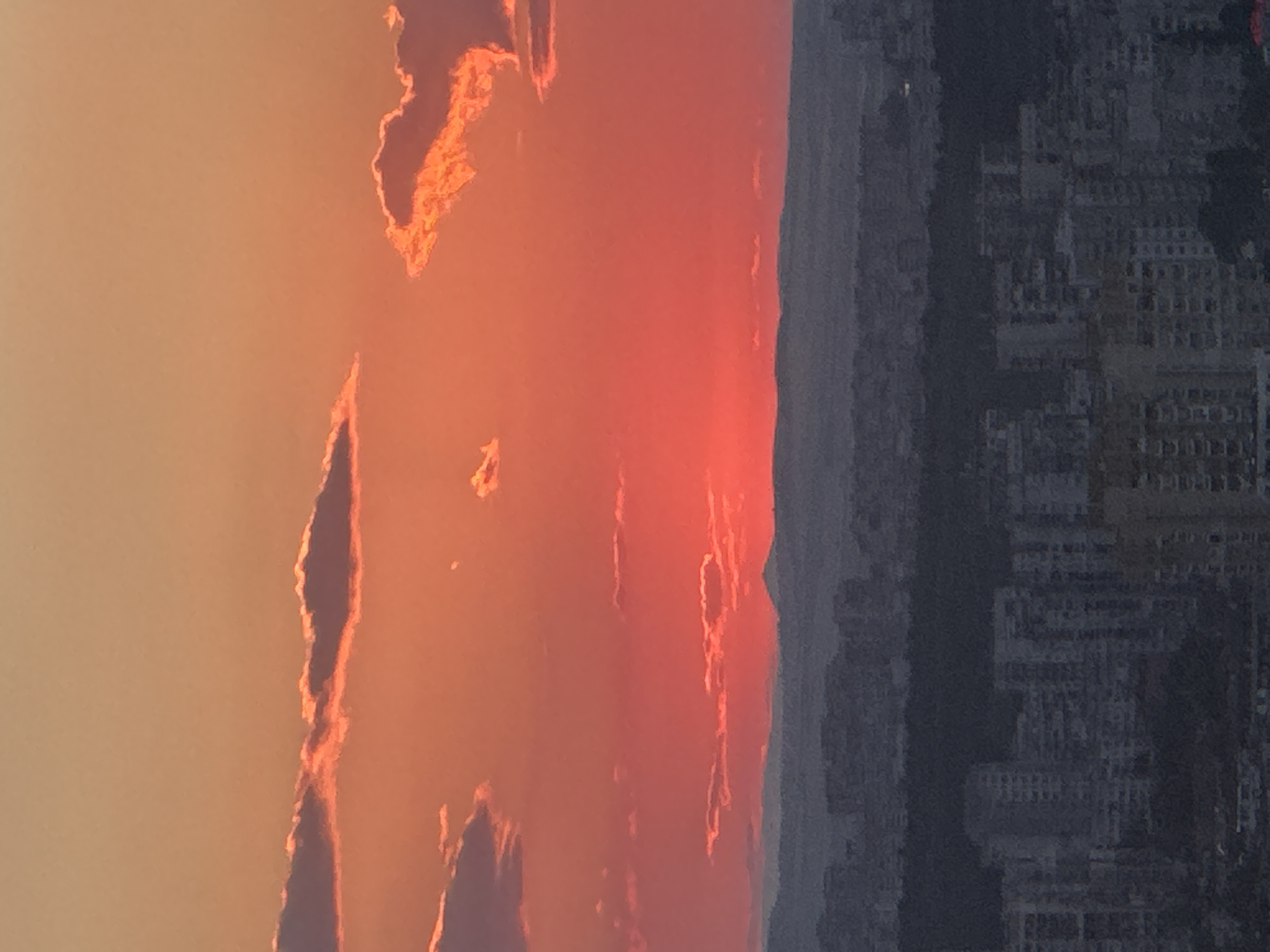 A high-angle, full shot captures a sunset over the city of Sofia, Bulgaria. The sky is a gradient of warm colors, transitioning from a pale orange at the top to a vibrant red near the horizon. Clouds are scattered across the sky, tinged with orange and pink from the setting sun. Below the sky, a silhouette of a distant mountain range can be seen, with a single peak visible. The middle ground features a dark, tree-covered hill, likely a park or a forested area within the city. The foreground is dominated by the urban landscape of Sofia, with numerous apartment buildings and structures rendered in shades of gray due to the low light conditions and distance. The buildings appear densely packed, characteristic of a city. No people or specific actions are discernible. The overall atmosphere is one of a tranquil evening in an urban setting, with nature's dramatic display of colors complementing the city's expanse.