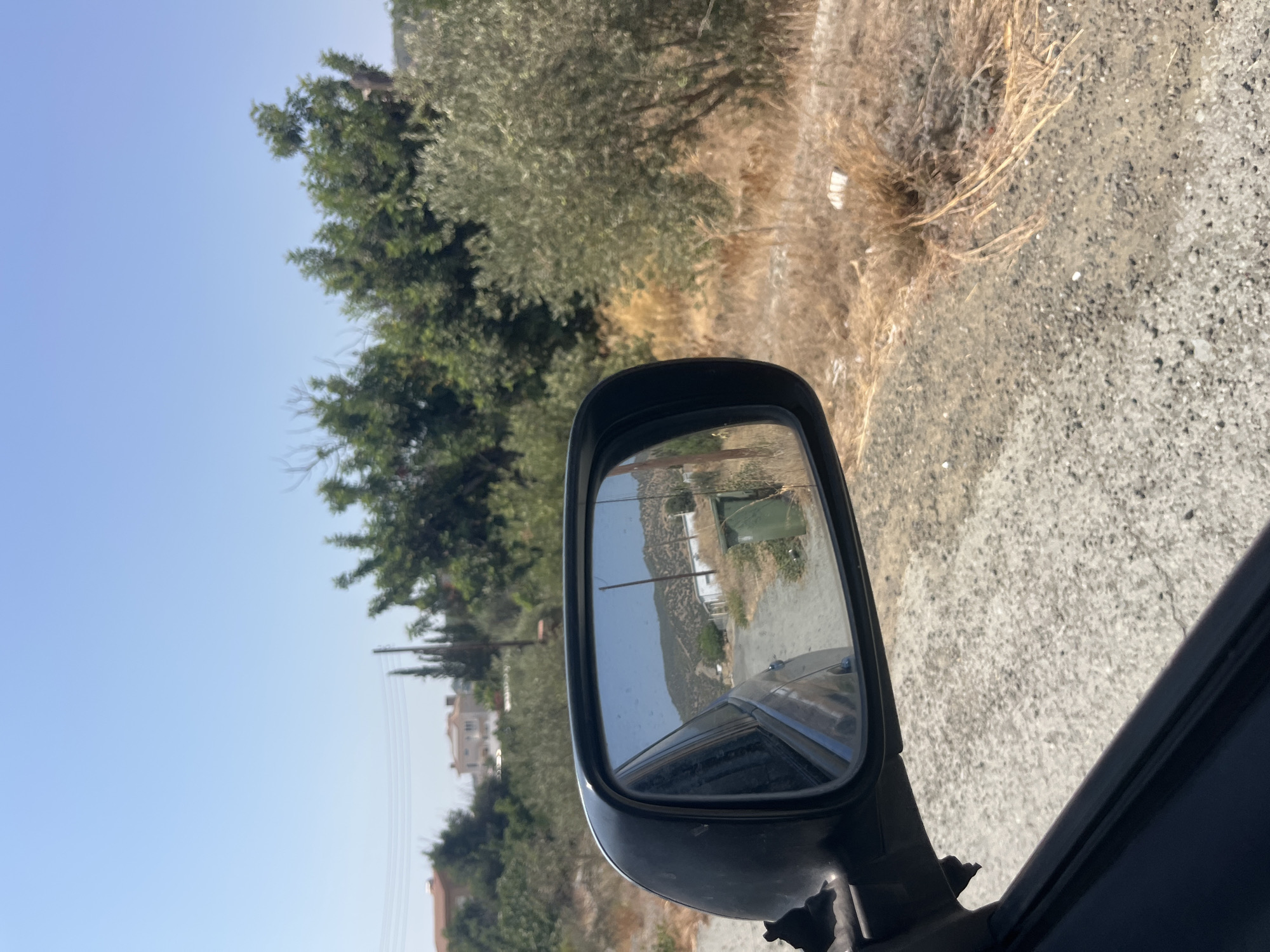 The image shows a view from a car in the village of Parekklisia, Cyprus. The scene is captured through a side-view mirror, reflecting a rural road. The road is unpaved and appears to be made of gravel and dirt. On either side of the road, there is dry, brown grass and scrub vegetation, indicating a dry climate.
In the reflection of the side-view mirror, the road curves slightly and is bordered by more dry vegetation and a few scattered buildings. There are some green bushes and trees visible in the distance, and a few residential buildings with tiled roofs can be seen on a gentle slope. A green trash bin is also visible on the right side of the road in the reflection.
Outside the mirror, the foreground shows the rough texture of the road surface. Beyond the road, to the right, are more dry, scrubby plants and some olive trees with silvery-green foliage. In the background, a clear blue sky dominates the upper portion of the image. The overall impression is of a warm, sunny day in a rural, Mediterranean setting. There are no people or animals visible in the image. The image composition suggests the car is moving forward along the road.