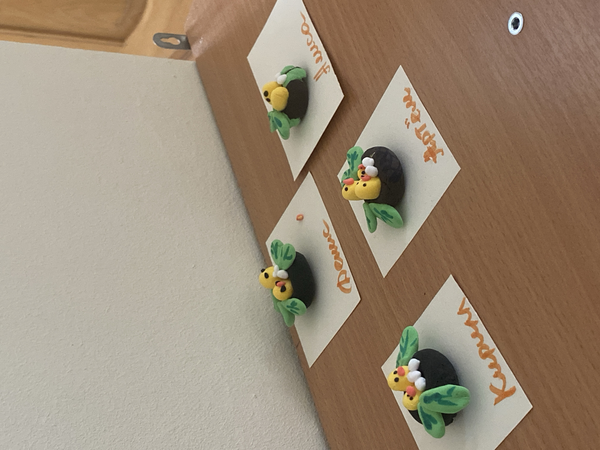 The image shows a wooden board affixed to a wall. The board has four diamond-shaped pieces of paper attached to it. Each paper has a small clay figure resembling a bug with green wings and yellow and white spots attached to it. Each paper also has a name written in orange ink. The names are "Luca", "Septem", "Denis", and "Kupsun". A metal eyelet is visible on the wooden board. The wall next to the board is white. The image was not taken in Sochi, Russia.
