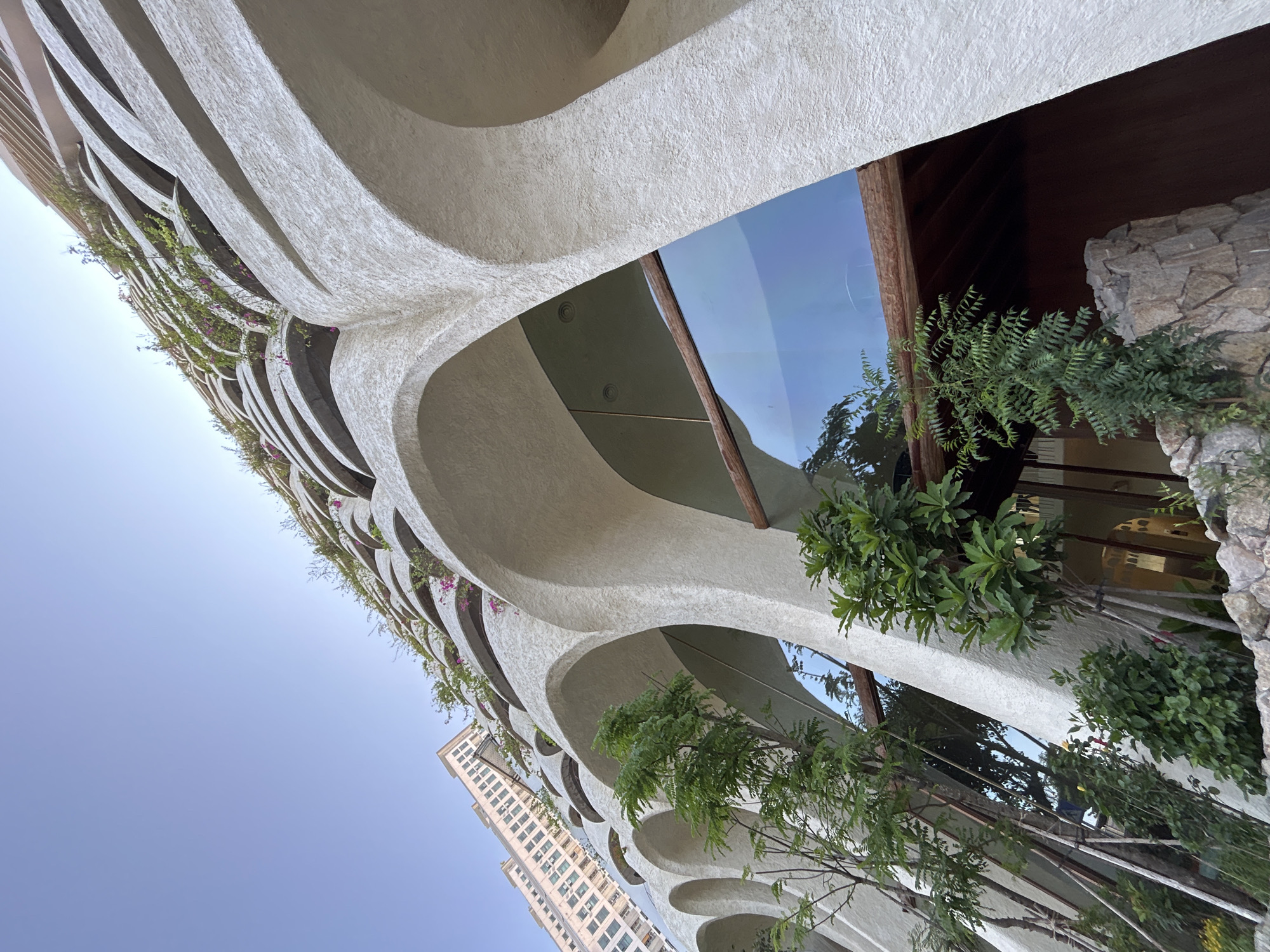 A low-angle shot captures the distinctive architectural features of a building in Bắc Nha Trang, Vietnam. The building's façade is characterized by curved, repetitive archways with rough white exteriors. Green foliage and small flowering plants are integrated into the arches. Glass panels fill some of the archways. Dark brown wooden structures and stone elements add contrast to the façade. The sky is visible in the gaps between the arches. A separate tall, rectangular building is situated in the background to the left of the frame. Some greenery is present in the foreground.