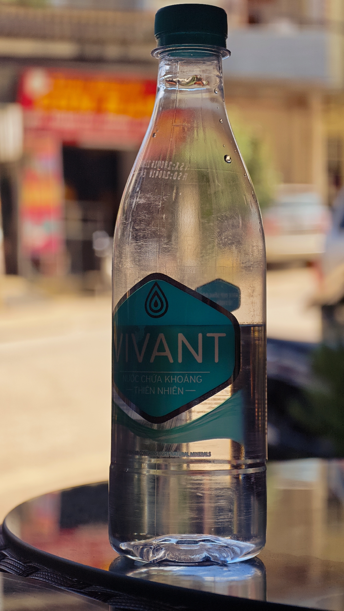 The image shows a partially consumed bottle of Vivant mineral water. The bottle is made of clear plastic with a green cap and a blue label. The label features a stylized water droplet and the word "VIVANT" in bold, white letters. Below that is Vietnamese text: "NƯỚC CHỨA KHOÁNG - THIÊN NHIÊN." Under that text is English text "WATER CONTAINS LIGHT NATURAL MINERALS". A few water droplets cling to the outside of the bottle. The bottle sits on a reflective surface, possibly a table.
The background is blurred, but suggests an outdoor setting, likely a street scene in Đồng Văn Commune, Vietnam. Hints of building facades in orange, red, and green can be seen, along with the vague outline of a vehicle. The lighting suggests a sunny day.