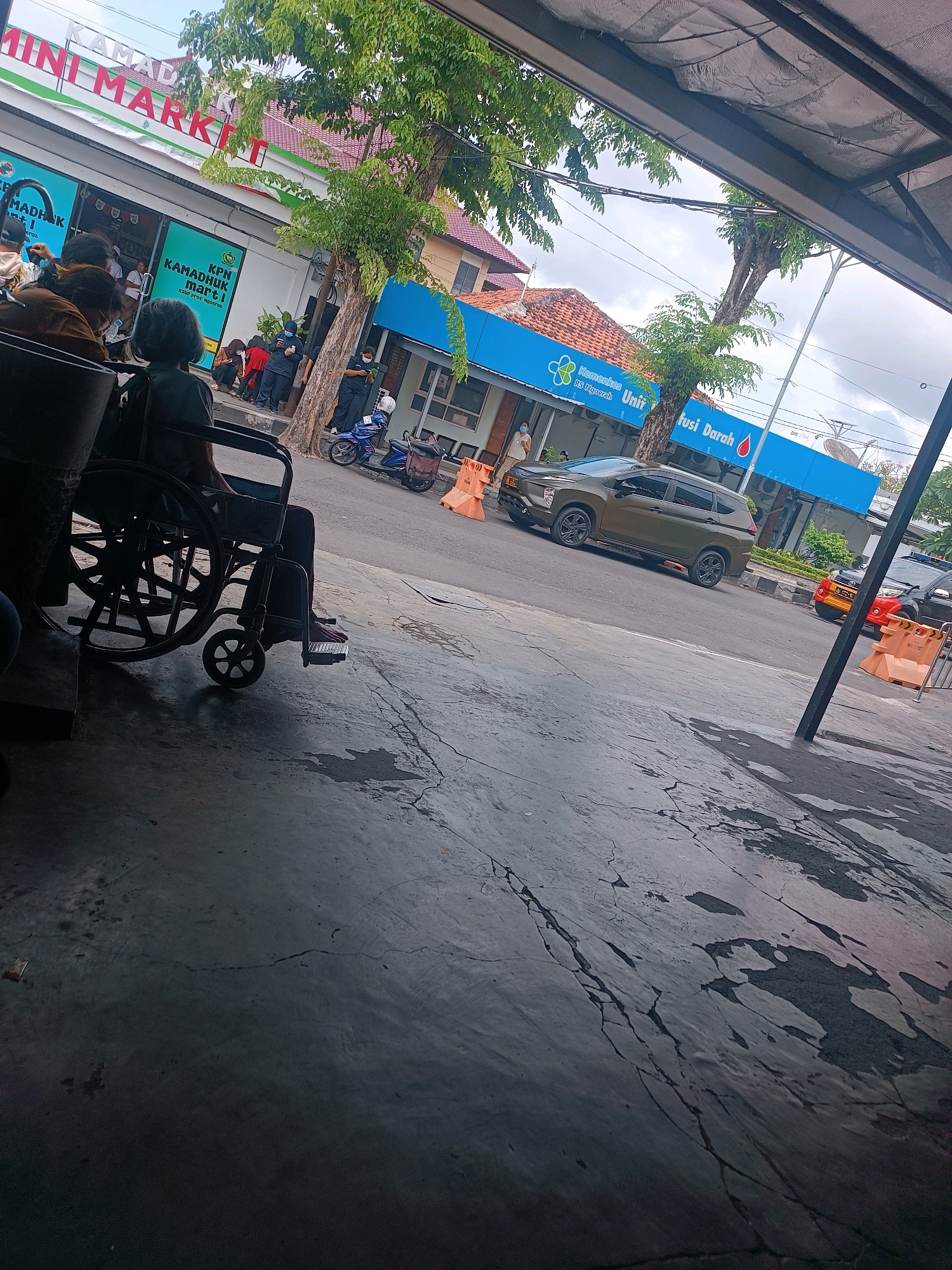 In Denpasar, Indonesia, outside a "MINI MARKET," a person sits in a wheelchair on a cracked concrete surface. Behind them, several people stand and sit. The MINI MARKET has green and white signage. Further down the street, a blue building with "Kemenkes RS. Ngerah Unit Fusi Darah" signage is visible. A dark green SUV and a red and white vehicle are parked on the asphalt road. The sky is cloudy.