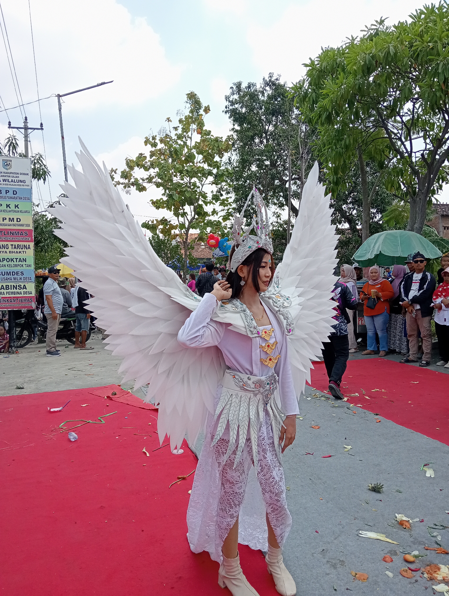 The image depicts a young woman in elaborate white costume, featuring large feathered wings and a crown, posing on a red carpet. She is on a street in Wilalung, Indonesia, indicated by signs that read "KECAMATAN GAJAH" and "DESA WILALUNG". The scene appears to be a public event or parade, with spectators visible in the background, some holding drinks and others dressed casually. A green umbrella is also visible, as are trees and a cloudy sky. The red carpet is scattered with debris, including leaves, papers, and what appear to be pieces of food. A motorcycle is parked on the left side of the image, and a few children are sitting on the ground. The woman's costume is detailed, with intricate silver and gold embellishments on her top, skirt, and crown. Her makeup is also elaborate, with eyeliner and glitter. The overall atmosphere seems festive, with a focus on the costumed individual.