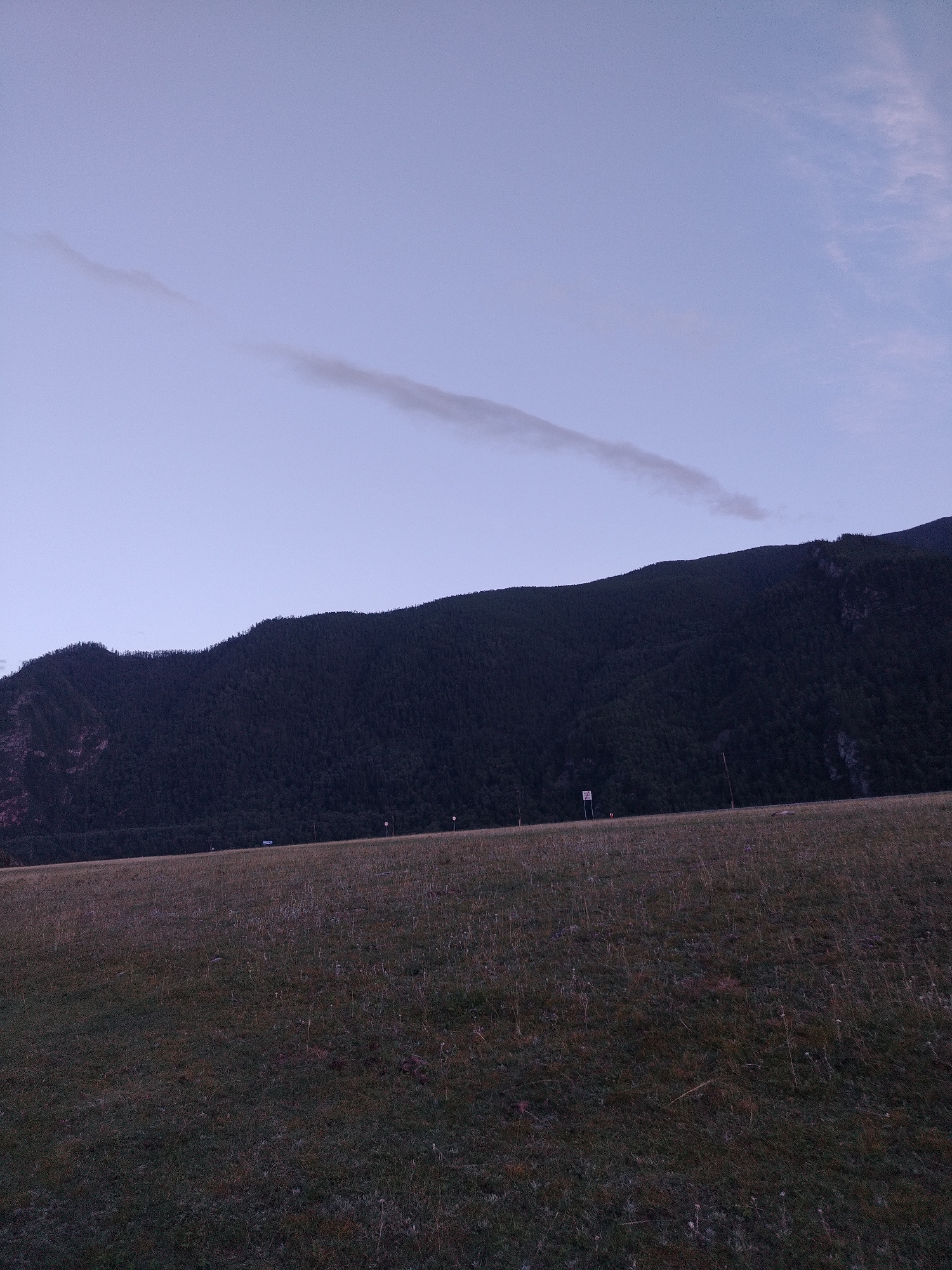 The image depicts a vast, low-lit grassland in front of a dark, tree-covered mountain range. The sky is a pale, dusky blue with a single wispy cloud stretching horizontally across the frame. The grassland appears dry and sparse, with hints of brown and green vegetation. In the distance, along the base of the mountain, faint signs of human activity are visible, including what appear to be utility poles and a small road or path. A white sign with some markings is also present near the road. The overall atmosphere is serene and natural, suggesting an outdoor setting in a rural or mountainous region. The location is identified as Iodro, Russia.