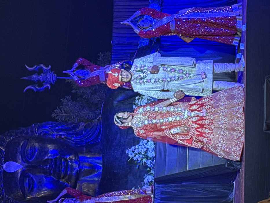 A staged wedding scene is lit with blue. The bride and groom, adorned in traditional red and white garments with floral garlands, are central. Flanking them are figures in similar red attire, positioned with hands in prayer. A large face sculpture, likely a deity, is prominent to the left, adjacent to a trident-wielding figure. The background features a mix of dark and blue hues.