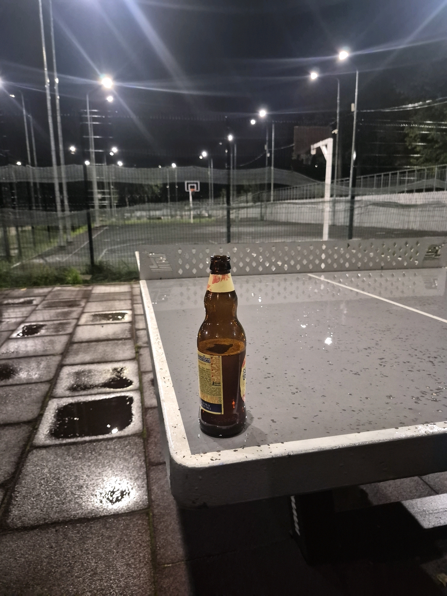 A brown glass beer bottle stands on a wet, grey outdoor ping pong table at night. The table is positioned on a paved area with square tiles and puddles, suggesting recent rain. In the background, a dimly lit outdoor sports court is visible, with a basketball hoop and backboard. Multiple tall light poles illuminate the area, casting long shadows and creating a slightly blurred effect due to the low light conditions. The scene appears to be located in an urban or recreational setting.
