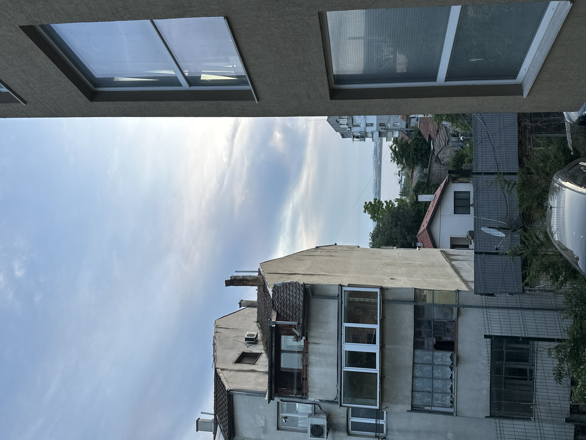 This media file is a photograph taken from a balcony, looking upwards and outwards towards a city scene. The foreground features the underside of a balcony with two windows. One window is clear, reflecting the sky, while the other has blinds.
Below the foreground balcony, an aged, multi-story building dominates the mid-ground. Its facade is a muted gray, with visible signs of wear and tear. Several windows are visible, some with clear glass, others with blinds or curtains. One section of the building features a glassed-in balcony area with multiple panes. Air conditioning units are mounted on the exterior walls, and a chimney protrudes from the roof. The roof itself is tiled with a mix of darker and lighter tiles.
Beyond this building, the scene opens up to a wider view of a cityscape. Various other buildings, some taller and more modern than the one in the mid-ground, are visible, along with trees and rooftops. In the distance, a body of water, likely the Black Sea, can be seen, with a hazy horizon. The sky is partly cloudy, with soft, wispy clouds against a pale blue backdrop. The lighting suggests it is daytime, possibly late afternoon or early morning, given the soft light.
The overall impression is one of urban life in Varna, Bulgaria, with a focus on the architecture and the expansive sky. There are no people visible in the image. The scene is quiet, with no obvious activity taking place. The details suggest a slightly weathered but lived-in environment. No visible text is present in the image.