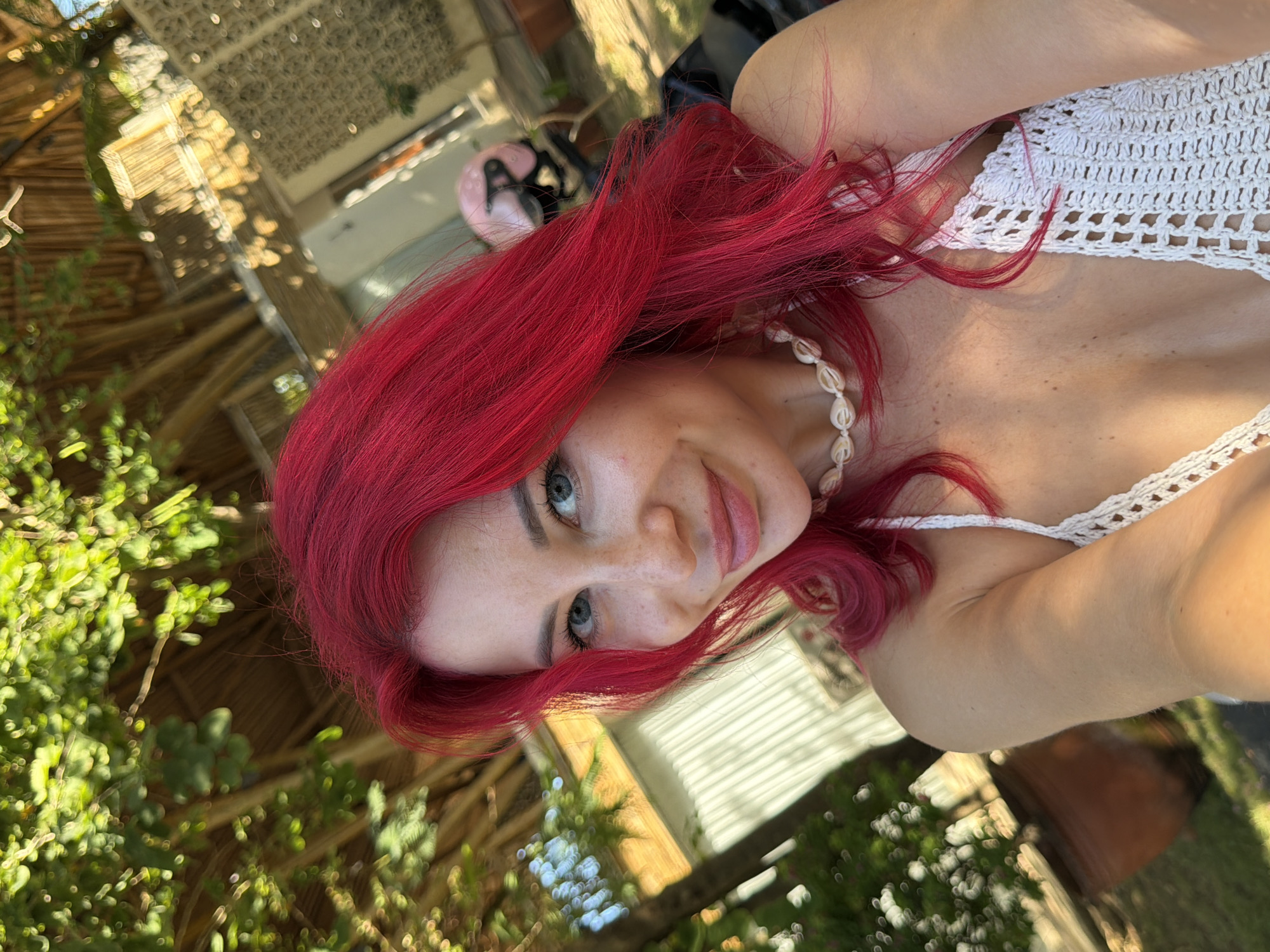 The image is a close-up selfie of a woman with vibrant red hair, wearing a white crocheted top and a seashell necklace. She is outdoors, with lush green foliage and a thatched roof structure visible in the background, suggesting a tropical or resort setting. The lighting indicates it is daytime, with sunlight filtering through the leaves. The woman has fair skin with freckles and blue eyes, and she is smiling at the camera. Behind her, partially visible, is a pink helmet resting on what appears to be a motorcycle or scooter. The overall scene evokes a relaxed, vacation atmosphere. While not explicitly visible, the described environment aligns with typical scenery found in Kuta, Indonesia, a popular tourist destination known for its beaches and resorts.