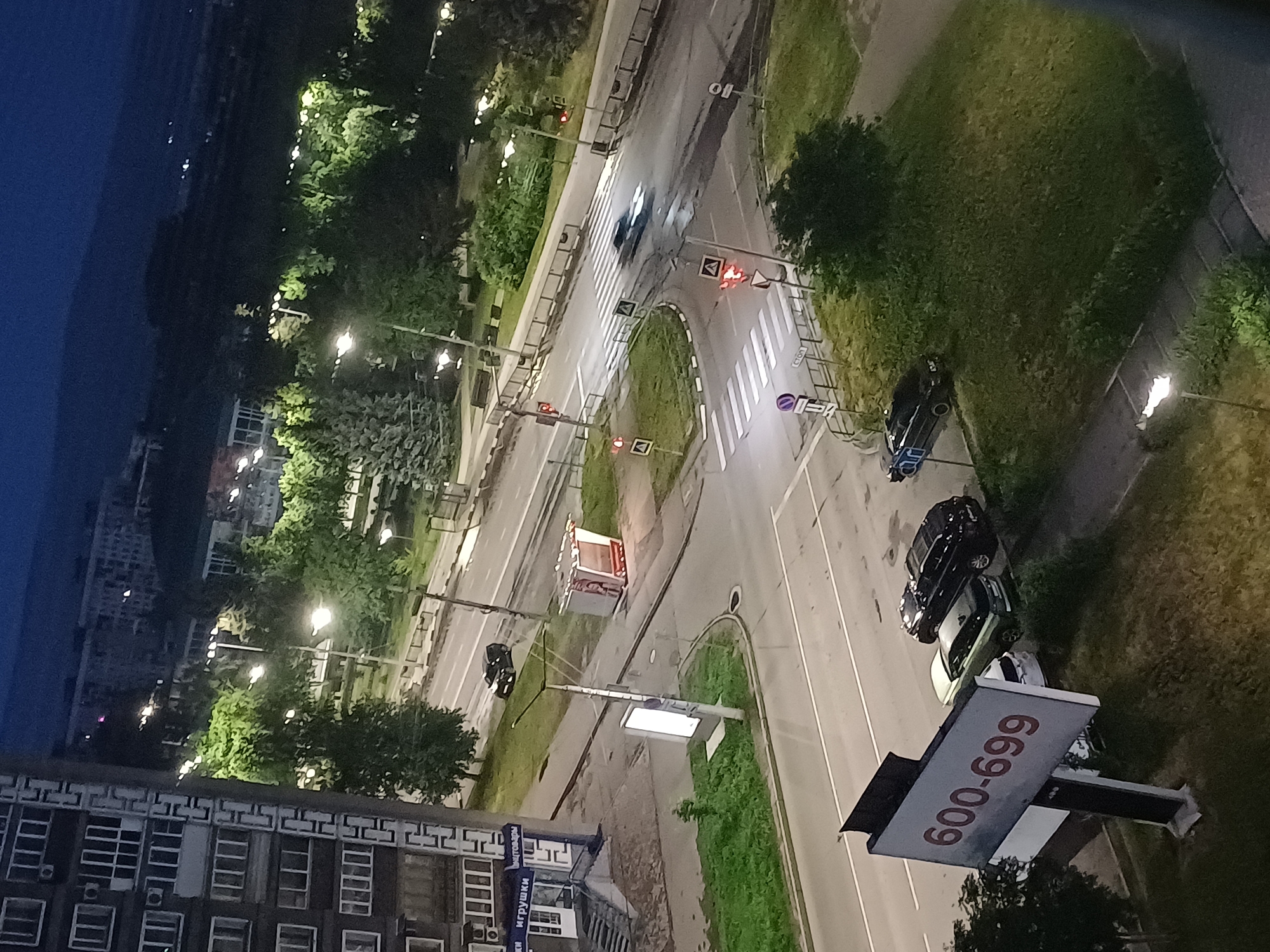 The image depicts a nighttime street scene in a city. In the foreground, a billboard displays the numbers "600-699". To the right of the billboard, three cars are parked: a black SUV, a white sedan, and a dark-colored sedan. A green area with bushes and trees is situated behind the parked cars. Further back, a road with a pedestrian crossing and traffic lights is visible. A car is seen in motion on the road, blurred due to movement. A bus stop shelter with a red bench is located on the side of the road. On the left side of the image, a multi-story apartment building with many windows is present. Beyond the street, more trees and buildings can be seen, with streetlights illuminating the area. The sky is dark, indicating nighttime.