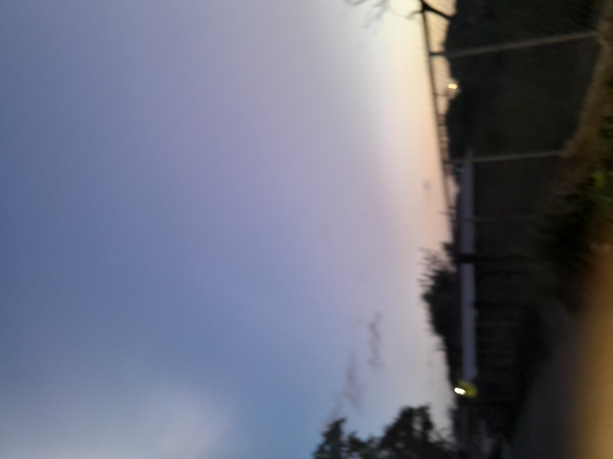 The image shows a blurry landscape at dusk or dawn. The sky transitions from a light blue at the top to a pale yellow near the horizon. A few wispy clouds are visible. In the foreground, there is a dark, out-of-focus area that suggests vegetation or ground cover. A chain-link fence runs horizontally across the middle ground, separating the foreground from the background. Behind the fence, the landscape is dark and indistinct, with what appears to be trees or bushes. There is a faint, out-of-focus circular light source visible above the fence in the mid-right of the image. The scene is likely a public area or the edge of a property, given the fence. The overall impression is one of a quiet, possibly rural or suburban setting. No specific landmarks or identifying features of Santa Ana, Venezuela, are discernible due to the blurriness and lack of detail.