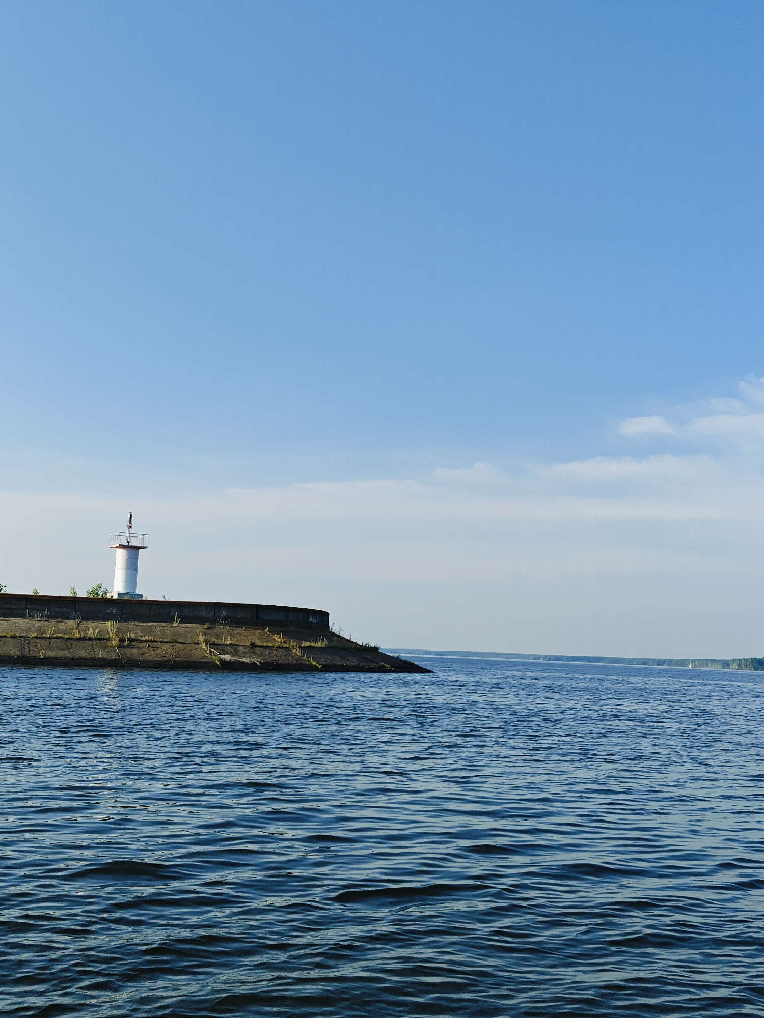 An outdoor, vertical, eye-level shot captures a serene view of a body of water, likely the Kama River, near the city of Chaikovsky, Russia. In the mid-left of the frame, a white lighthouse with a red top stands atop a curved, concrete embankment. The embankment is covered with sparse, dry vegetation. The water in the foreground exhibits small ripples and is a deep blue. In the distance, a faint, tree-lined shore is visible, with what appears to be a small sailboat on the horizon. The sky is a clear, light blue with a few wispy white clouds.