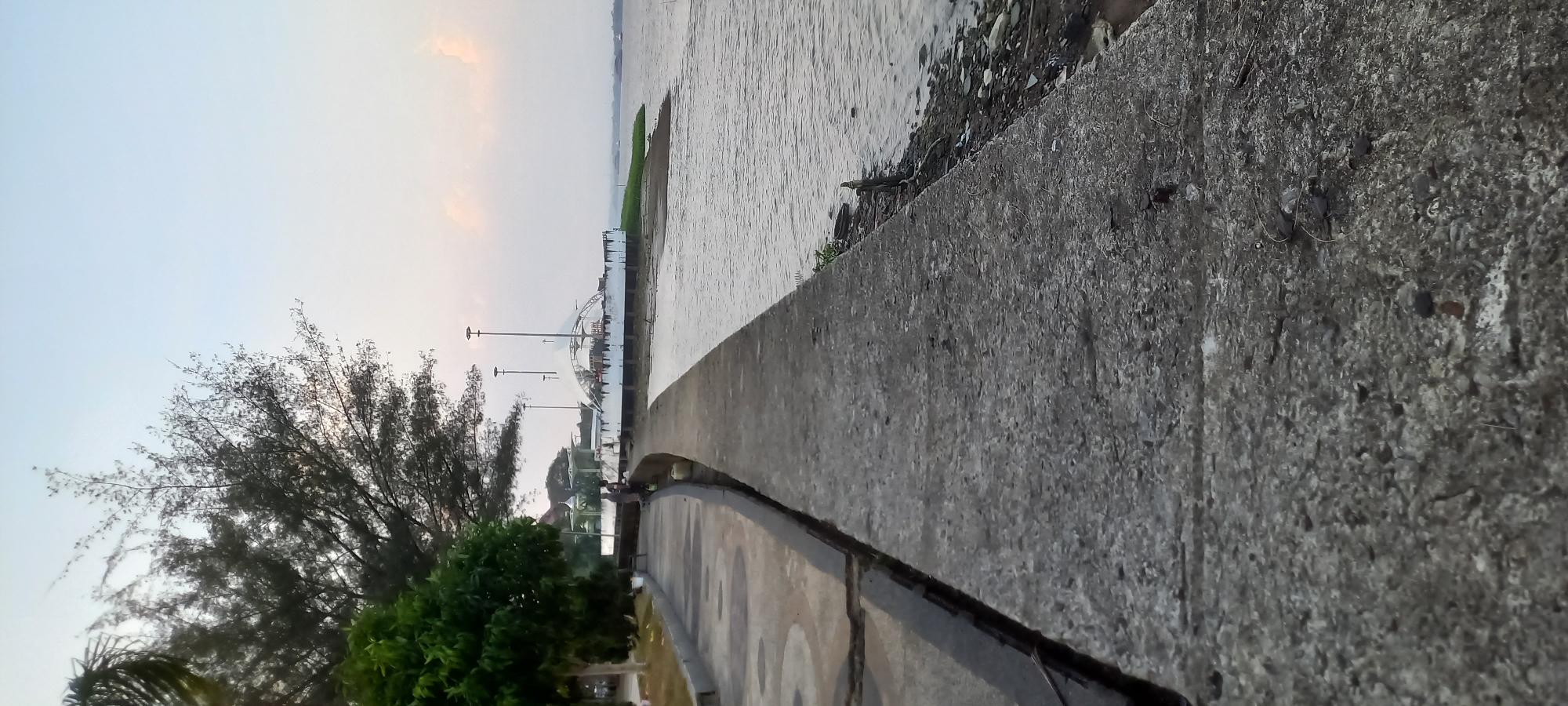The image depicts a waterfront promenade in the city of Melayu, Indonesia. In the foreground, a cracked concrete walkway runs along the edge of a body of water. To the left of the walkway, a tree-lined park area is visible, with a few people walking or standing. In the background, a white, curved structure with a canopy and multiple light poles stands near the water. The sky above is overcast with a soft, diffused light. The water itself appears calm, with some debris visible along the shoreline. The scene suggests a public recreational area, likely a park or a waterfront plaza.
