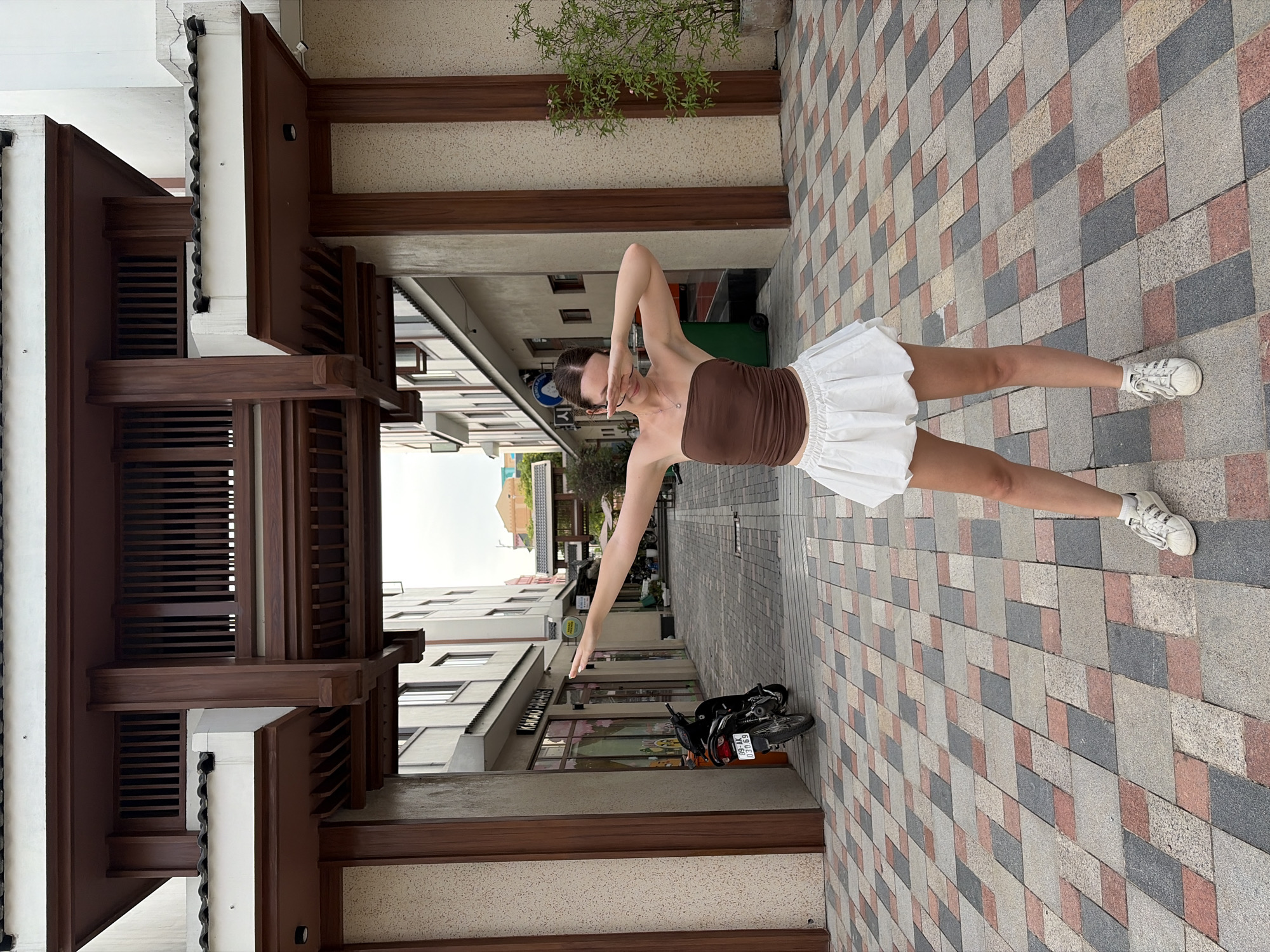 A young woman stands in what appears to be a commercial alleyway in Nghĩa Trụ Commune, Vietnam. She is wearing a brown strapless top and a white bubble skirt, with white sneakers. The woman is posing with her arms extended, her hands forming a V-shape over her face, a gesture often associated with "dabbing". Behind her, a paved walkway is laid with a patterned arrangement of rectangular tiles in shades of grey, red, and beige. The alleyway is flanked by buildings with light-colored facades and dark wooden architectural elements. To the left, a black motorcycle is parked, and a shop front with the text "KAKAO FRIENDS" is visible. To the right, a potted plant with green foliage adds a touch of nature to the urban setting. The overall impression is one of a bright, outdoor pedestrian area, likely a shopping or entertainment district.