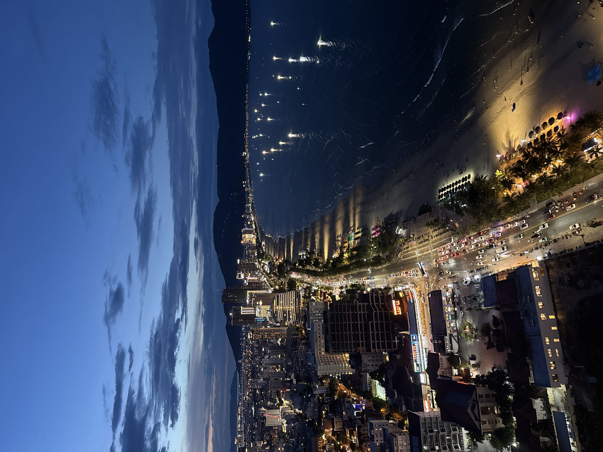 An aerial, landscape-oriented photograph captures the vibrant cityscape of Da Nang, Vietnam, at dusk or early night. The sky, a deep twilight blue with scattered clouds, fades into the dark silhouettes of mountains on the horizon.
To the right, the dark expanse of the ocean is illuminated by numerous bright lights, likely from fishing boats or vessels, creating shimmering reflections on the water. A wide sandy beach stretches along the coastline, brightly lit by artificial lights that cast long shadows of palm trees and beach amenities onto the sand. Scattered individuals can be seen walking along the shore.
Adjacent to the beach runs a busy, multi-lane coastal road, bustling with traffic. Headlights and taillights from numerous cars and motorbikes form streaks of light, indicating continuous movement. Beyond the road, the city sprawls inland with a dense network of buildings, including high-rises and commercial establishments, all brightly lit. The visible text "LANG CA" appears on signage on multiple buildings along the main road. The scene emanates a lively urban energy typical of a thriving coastal city transitioning from day to night.