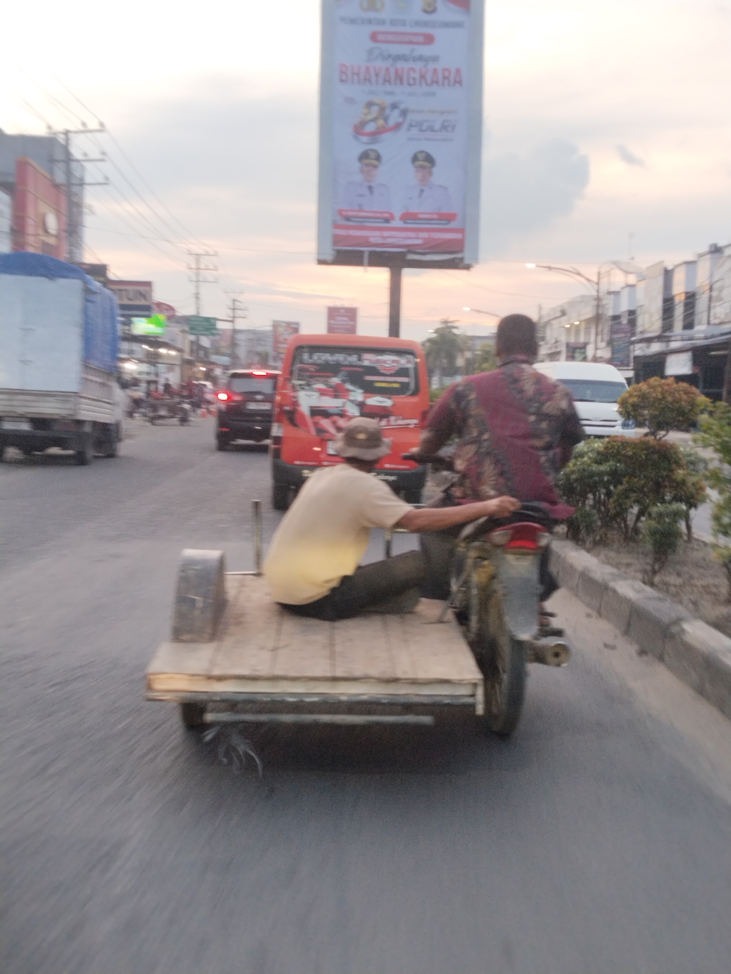 The image captures a street scene in Lhokseumawe, Indonesia, likely during daylight hours given the lighting. In the foreground, a person wearing a light-colored shirt and a cap is seated on a wooden trailer attached to a motorcycle. The trailer appears to be made of weathered wood and has metal framing. The motorcycle, ridden by another person wearing a patterned shirt, is in motion, indicated by the motion blur. The motorcycle itself is a common model with a visible rear wheel and exhaust pipe.
The street is paved and appears to be a busy road with other vehicles present. To the left, a large white truck with a blue tarp covering its load is partially visible. Further down the street, an orange vehicle, possibly a truck or van, is seen with its brake lights illuminated, suggesting traffic. A white van is also visible in the middle ground.
Along the right side of the road, there is a concrete curb and a patch of vegetation with dense bushes. In the background, a large billboard displays promotional content, featuring images of two individuals in uniform and text in Indonesian, including the word "POLRI" (Indonesian National Police). The urban environment is evident with buildings and utility poles with numerous wires. The sky is overcast with soft, diffused light. The overall impression is that of everyday urban traffic and commerce in Lhokseumawe.