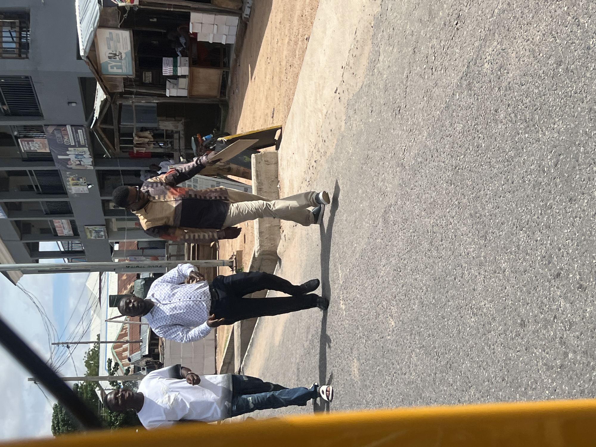 The image displays a daytime street scene in Jalingo, Nigeria. Three adult males are walking from left to right across the foreground on an asphalt road. A yellow vertical element partially frames the left side of the image, suggesting a view from within a structure or vehicle.
From left to right:
The first man wears a white short-sleeved t-shirt with a dark graphic design on the chest, faded blue jeans, and white slide sandals. He is smiling and appears to be looking towards the man in the center. His left arm is bent, hand partially visible.
The second man wears a light-colored, long-sleeved button-down shirt with a repeating pattern, dark trousers, and dark dress shoes. He maintains a neutral expression and looks forward. His arms are slightly bent at the elbows.
The third man wears a long-sleeved collared shirt featuring a multi-colored abstract or geometric pattern, light-colored trousers, and dark sneakers with white soles. He holds a flat, light-colored rectangular object, possibly made of wood or cardboard, in his right hand, extended downwards. He looks slightly towards the ground and his right.
The road surface is a grey asphalt with visible aggregate. To the right of the road, there is a raised concrete curb, followed by a concrete sidewalk, and then an unpaved dirt shoulder. The background features various buildings under a bright, partly cloudy sky. A multi-story grey building with black railings and balconies is prominent on the right, displaying banners, one of which reads "CELEBRITY COSMETICS". In front of this building, closer to the street, are smaller, makeshift commercial stalls with corrugated metal roofs. One stall has a sign with the letters "IFY" clearly visible, and stacks of white boxes are visible near its counter. A dark motorcycle is parked between the main building and these stalls. Utility poles and a network of overhead wires are visible in the left-mid background, above other structures with corrugated roofs. The scene is well-lit by direct sunlight, casting distinct shadows of the individuals and objects on the road and ground.