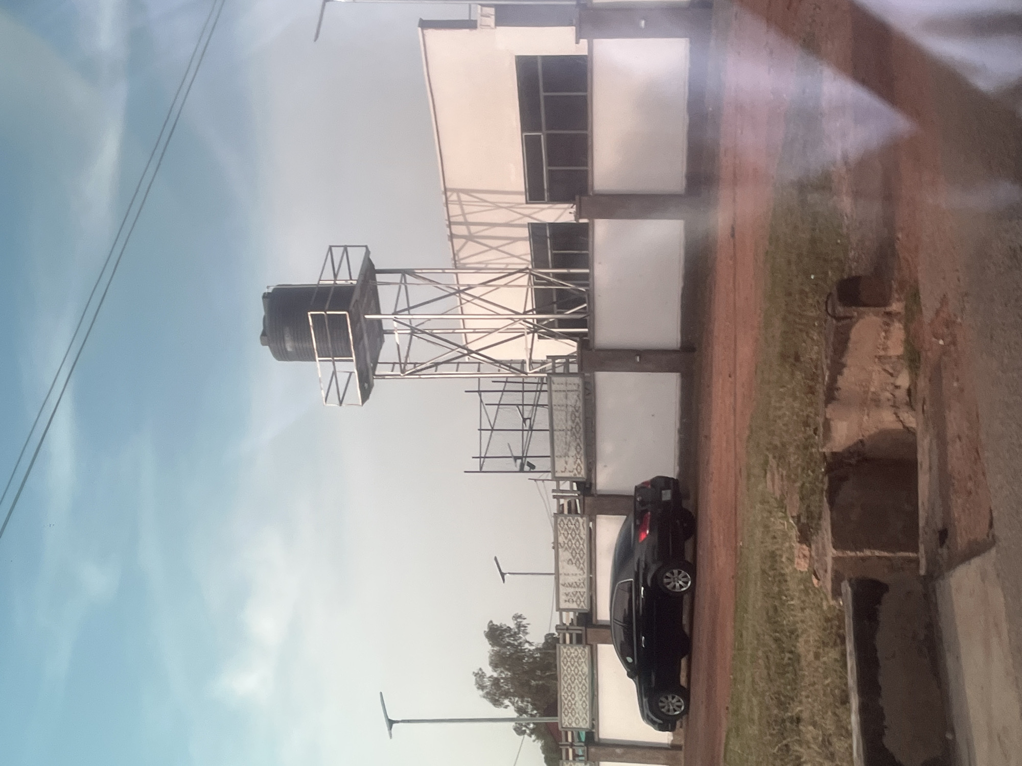 This is a vertical, full shot of an outdoor scene that appears to be in Jalingo, Nigeria. The image features a water tower with a large black tank supported by a metal frame. Adjacent to the water tower is a modern-looking building with a white facade and multiple windows. A dark-colored sedan is parked near the building. The ground is a mix of dirt and grass. Overhead power lines are visible against a pale blue sky with some clouds, suggesting it might be daytime, possibly late afternoon due to the soft lighting. There are also streetlights present. The scene suggests a functional area, perhaps a residential or commercial complex. The image is slightly blurry and has a reflective sheen, indicating it might have been taken through a window or with a camera that has a strong glare. No people are visible in the frame.