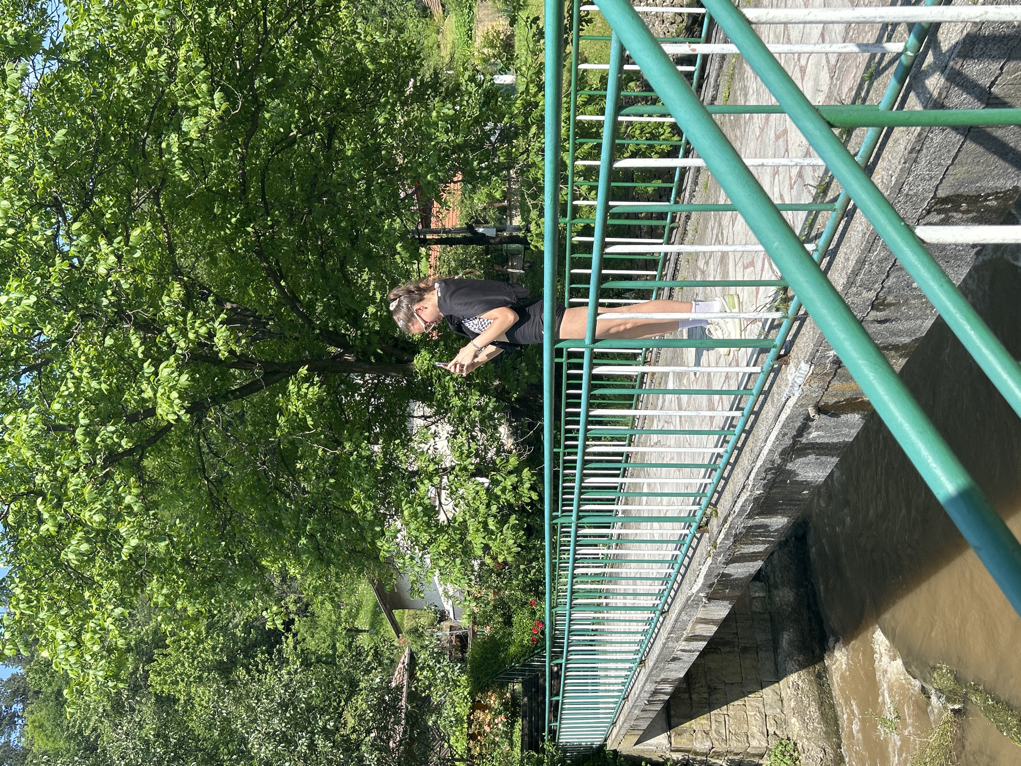 The image captures a vibrant outdoor scene, likely a rural or village setting within an unknown city. A young female individual stands on a concrete bridge that spans a muddy brown river or stream.
The central figure, a female with light brown hair tied in a high ponytail, wears glasses with light-colored frames. She is dressed in a black short-sleeved top, a black and white checkered sports bra, black shorts, white socks, and light-colored athletic shoes (possibly light green or yellow). She stands facing slightly left from the viewer's perspective, holding a smartphone with both hands and appears to be operating it, possibly taking a photo or video.
The bridge features a concrete deck paved with light-colored, patterned tiles. Its railing is composed of alternating green and white vertical metal bars, topped by a darker green horizontal handrail. The river beneath the bridge flows with visibly turbid, brownish water.
The background is dominated by lush, dense green foliage from numerous trees, indicating a heavily vegetated environment. Several houses with white walls and reddish-brown roofs are partially visible through the trees on the far side of the river, suggesting a small settlement or scattered homes. Steps and some flowering plants (including red roses) are also visible near one of the houses. The scene is brightly lit by sunlight, casting distinct shadows, particularly under the bridge and from the railings. A clear blue sky is visible in the upper right corner.