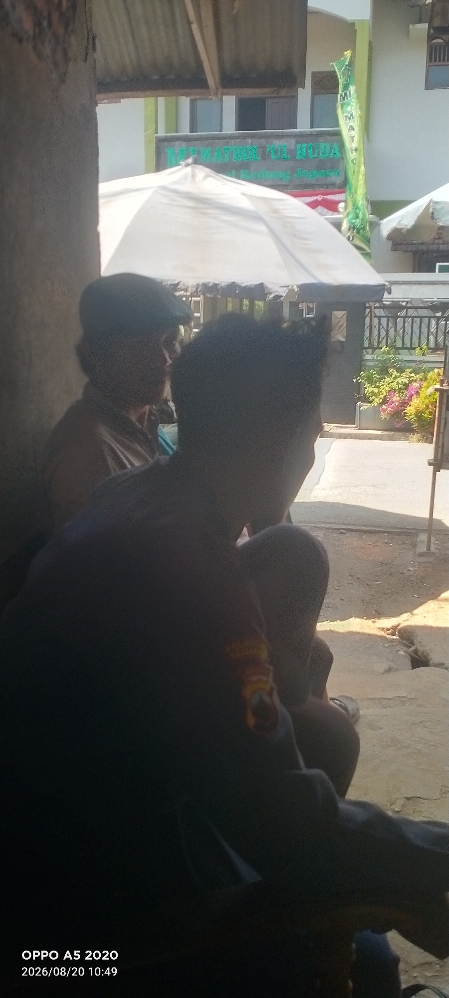 The image depicts an outdoor scene with two individuals sitting. The person on the left is wearing a cap and a light-colored shirt, and appears to be looking to the right. The person on the right is mostly in silhouette, wearing a dark shirt with an emblem on the sleeve. They are positioned to the right and slightly in front of the person on the left. In the background, a building with a green sign that reads "MADRASAH MATHOL'UL HUDA" is visible. A white umbrella or canopy is positioned between the foreground figures and the building. The ground appears to be a dirt or paved surface with some debris. The time stamp on the image indicates "OPPO A5 2020 2026/08/20 10:49," suggesting the photo was taken in Bugel, Indonesia, on August 20, 2026, at 10:49 AM. The lighting suggests it is daytime, with sunlight casting shadows.