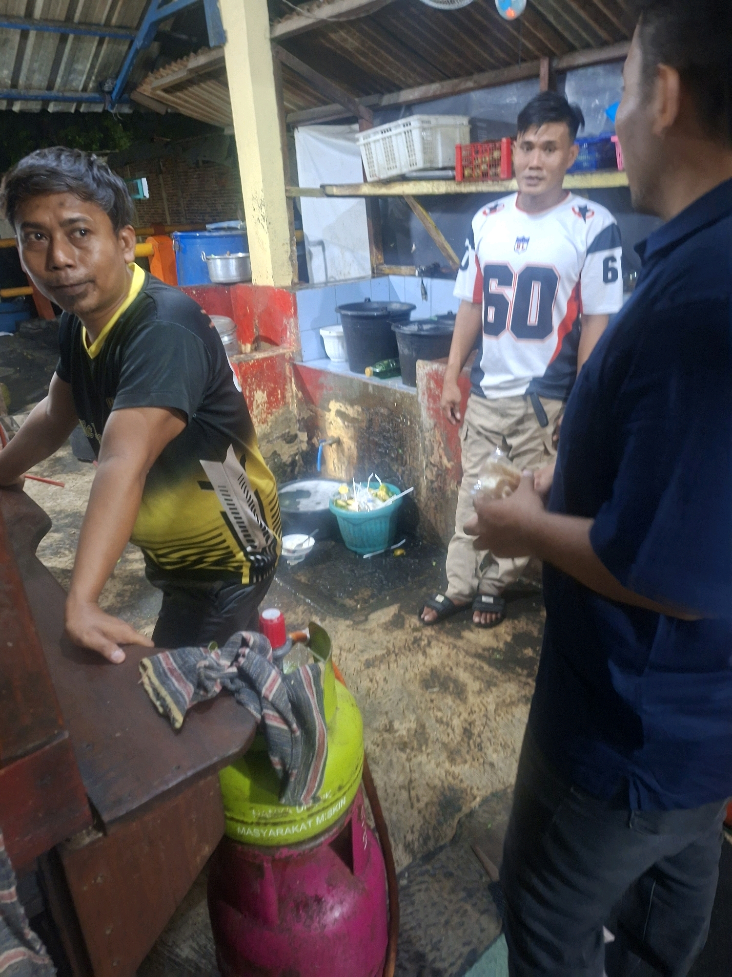 The image depicts an outdoor scene, possibly a food stall or market, in Semarang, Indonesia. In the foreground, a man wearing a black and yellow t-shirt with a geometric design stands leaning on a wooden counter. His left hand rests on the counter, and his right arm is bent. To his right, a large green gas cylinder with pink base is visible, partially covered by a striped cloth.
Behind the first man, another man is seen wearing a white t-shirt with the number "60" in large black numerals and a navy blue collar and sleeves. He is wearing light brown cargo pants and black sandals. He appears to be talking to another individual, whose upper body is visible on the far right of the image. This third person is wearing a navy blue shirt and dark pants, and is holding a clear plastic bag containing what appears to be food.
The environment includes a wooden roof structure overhead supported by a yellow pillar on the left. In the background, there are shelves with various items, including white crates, red plastic containers, and black buckets. A blue container and a metal pot are also visible. The ground is concrete and appears wet, suggesting recent rain or cleaning. The overall lighting is dim, indicating it might be evening or under artificial lights. The scene suggests a casual, everyday interaction among people in a bustling environment.