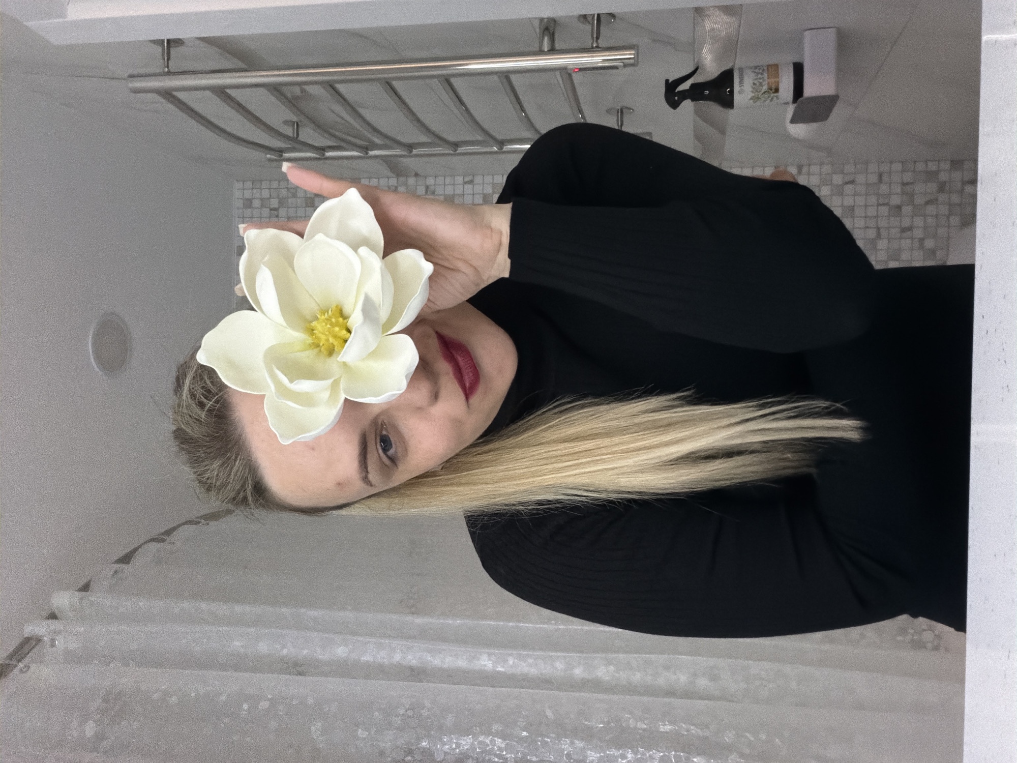 The image is a sideways portrait of a woman indoors, rotated 90 degrees clockwise. The woman, with blonde hair and fair skin, is wearing a black long-sleeved top. She holds a large, white artificial flower, resembling a magnolia, in front of her face, partially obscuring her left eye and forehead. Her visible features include a blue or gray eye, a defined eyebrow, and bright red lipstick. Her long blonde hair cascades to the left side of the frame.
The background suggests a bathroom or utility area, featuring light-colored elements. Behind her head and shoulder, a silver multi-tiered towel rack or shelf is mounted on a light-colored tiled wall composed of small, square mosaic-like tiles in shades of gray and white. On a surface further to the right, a dark-colored spray bottle with an illegible label and a white rectangular dispenser are visible. The lower left portion of the image shows a textured, translucent white shower curtain. The overall lighting is soft and even, indicating an indoor setting with artificial illumination. The woman's expression is neutral, and her pose indicates she is intentionally presenting the flower to the camera.