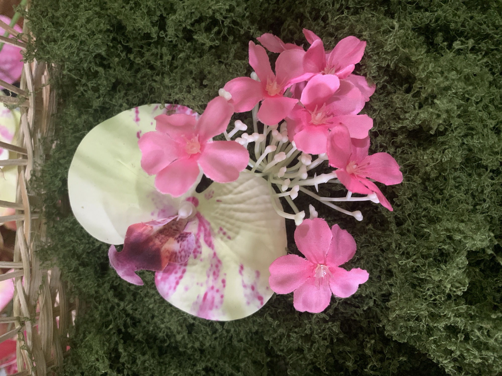A close-up, overhead shot shows an arrangement of artificial flowers on a bed of green moss. A large, pale yellow orchid with pink speckles and a deep pink center sits behind several bright pink, five-petaled flowers. A cluster of small, white, bud-like structures is interspersed with the pink flowers. The flowers are placed on a textured green surface that resembles moss. In the upper background, a woven basket is partially visible. The image is brightly lit, with shadows cast by the flowers onto the moss. The location in Kuala Pembuang, Indonesia, is not discernible from the visual elements of the image.