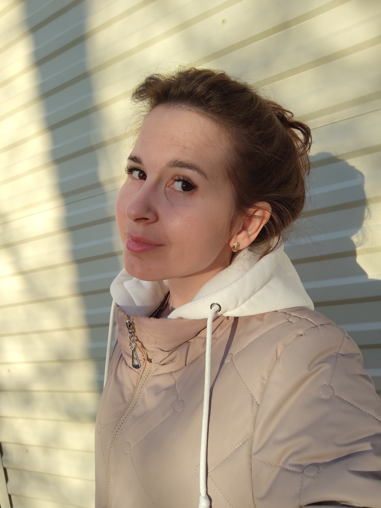A young woman is captured in a close-up outdoor portrait, looking over her left shoulder towards the viewer with a slight, closed-mouth smile or smirk. Her head is tilted, and her dark brown eyes with prominent eyelashes engage directly with the camera. She has light brown hair, styled casually with some wisps visible around her face, and wears small gold hoop earrings.
She is dressed in a light beige or pink quilted jacket, possibly a puffer style, layered over a white hooded sweatshirt with white drawstrings. The jacket features a metallic silver zipper and quilted stitching forming diamond patterns and circular indentations.
The background consists of a light-colored (likely white or very light beige) building wall with horizontal siding. Strong, elongated shadows, presumably cast by the woman and her surroundings, fall diagonally across the wall. This prominent shadowing suggests direct sunlight from a low angle, indicating either early morning or late afternoon. The jacket implies that the weather is cool or cold. The overall expression on the woman's face conveys a sense of playfulness or confidence. The location is Фоки, Russia. No text is visible in the image.