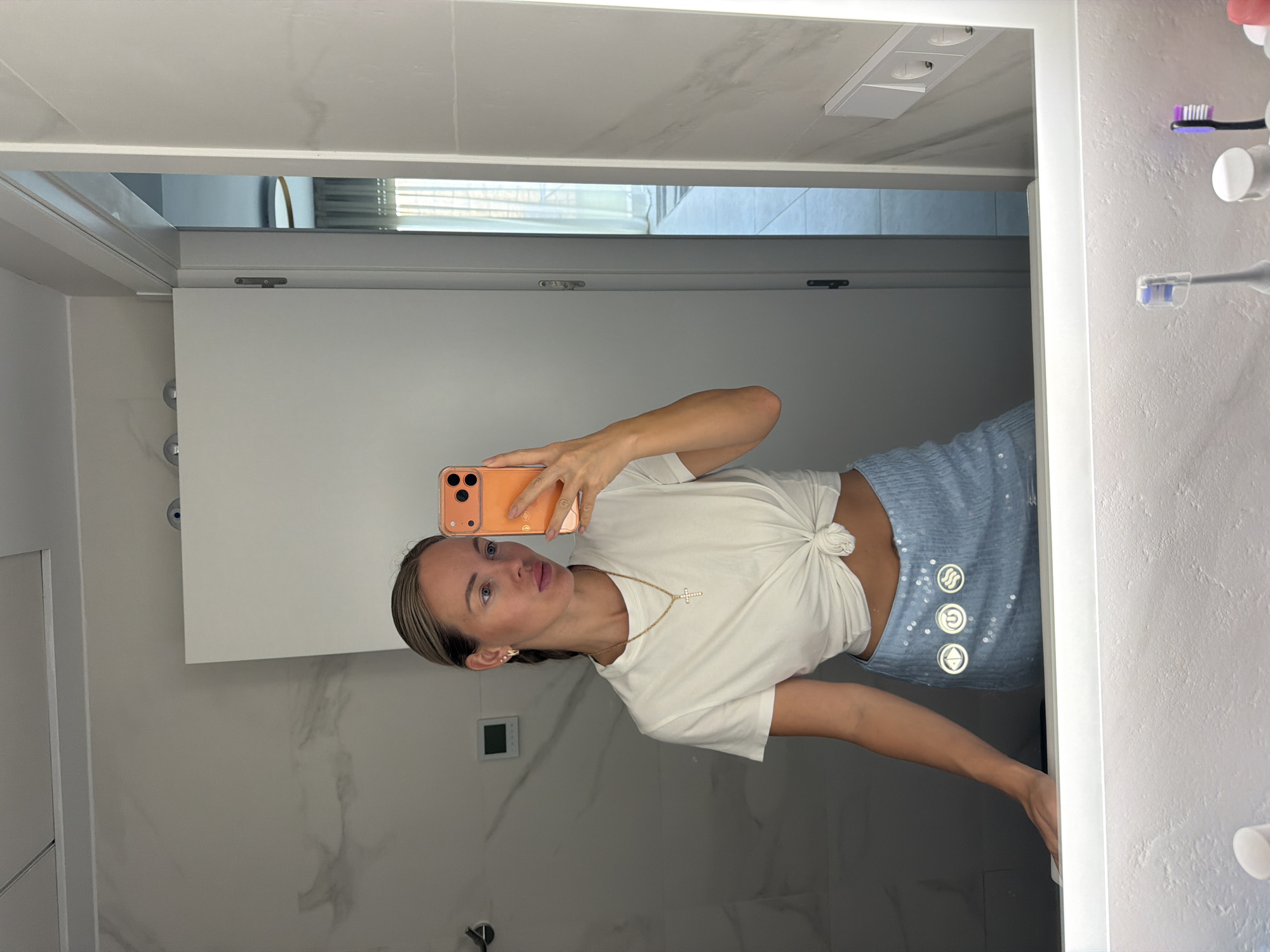 The image depicts a woman taking a mirror selfie in a modern bathroom. She is wearing a white knotted t-shirt and a light blue sequined skirt. She is holding an orange smartphone to take the picture. The bathroom features marble-patterned walls, a large illuminated mirror, and various toiletries in the foreground. In the background, a door is visible, leading to a well-lit room with curtains. The woman's hair is pulled back in a braid, and she is wearing a delicate necklace. The overall scene suggests a private, domestic setting, possibly in a hotel or residential property. The city of Finestrat, Spain, is not explicitly visible or indicated within the image itself.