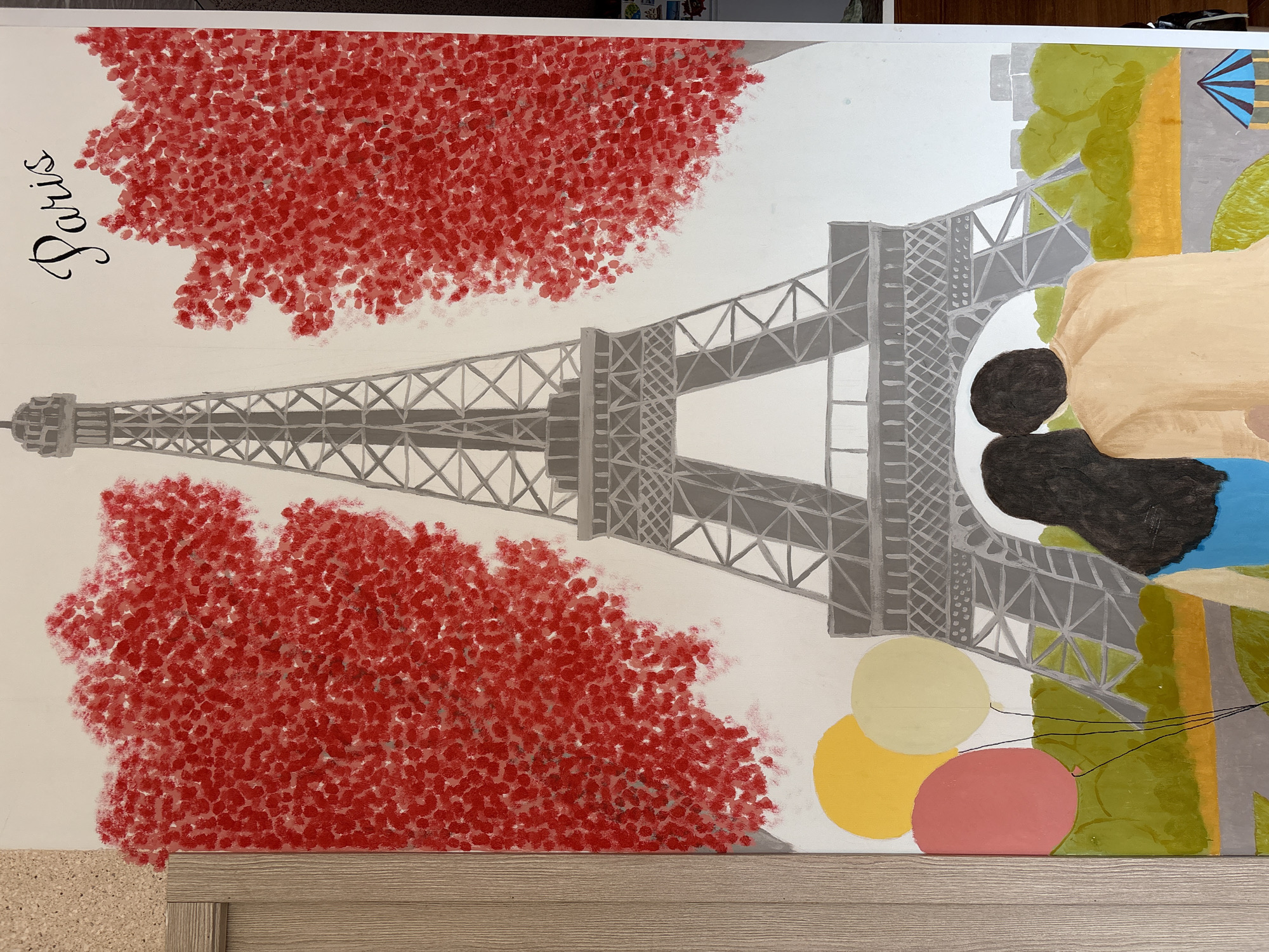 This is a painting depicting the Eiffel Tower in Paris, France. The painting features the iconic tower in a grey monochrome style. On either side of the tower, there are large, abstract trees rendered in red dotted brushstrokes, suggesting a romantic or autumnal setting. The sky is white. In the foreground, a couple is seen from behind, looking towards the Eiffel Tower. They are positioned in the center of the painting. The woman has dark hair and is wearing a blue top, while the man has dark hair and is wearing a tan shirt. To the left of the couple, three balloons are depicted in pink, yellow, and off-white. The ground below is painted with green foliage and an orange area, with a grey path. In the far background, there are faint outlines of buildings. The word "Paris" is written in cursive script in black ink at the top right of the painting. The painting is hung on a wall. There is no information in the image to suggest a location in the city of Ветлужский, Russia.