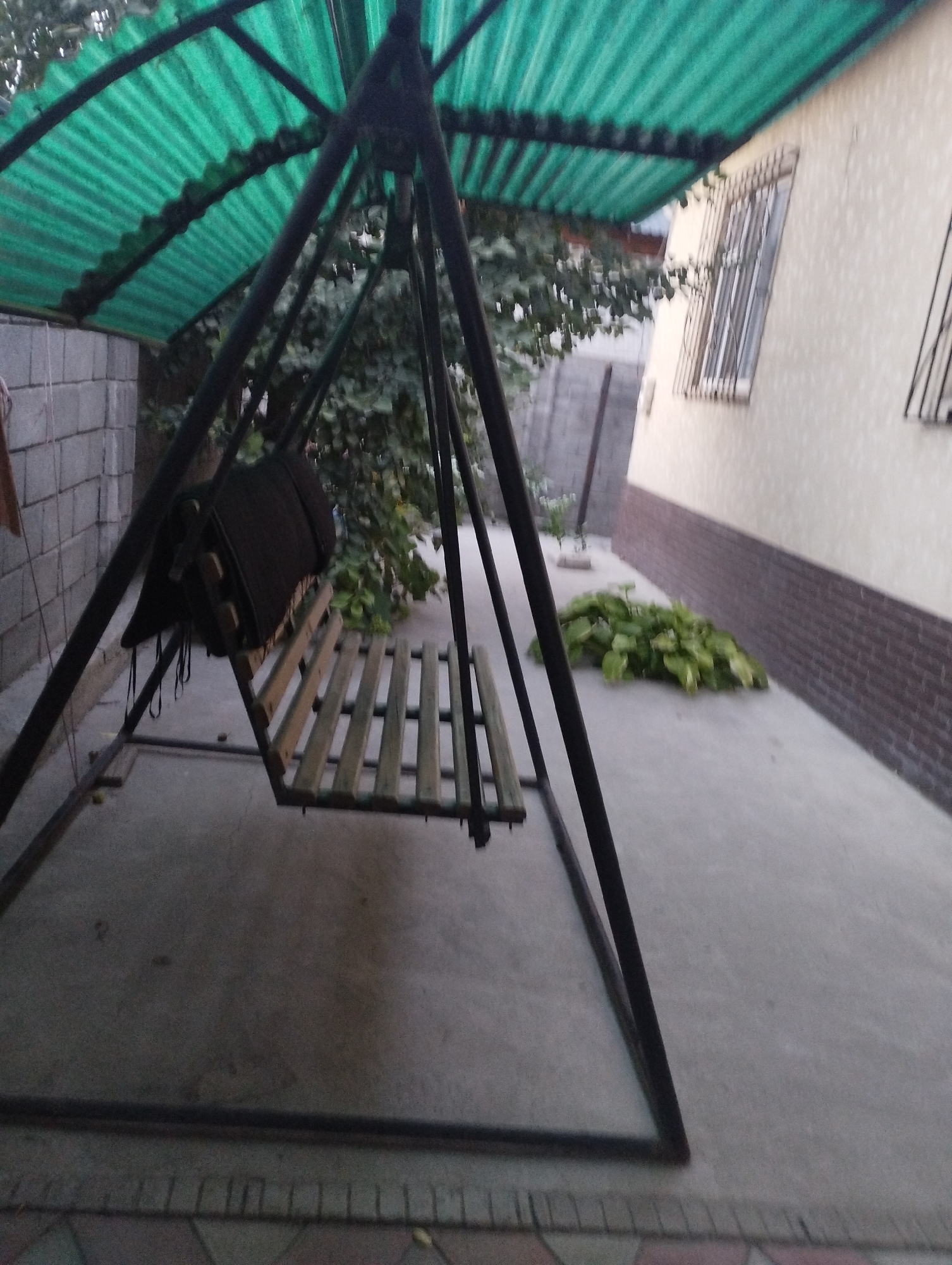 A static, eye-level, outdoor shot captures a metal swing set with a green corrugated plastic roof. The swing seat is made of wooden slats and a dark cushion is draped over the backrest. To the left of the swing, a grey cinder block wall is visible, and to the right, a light-colored building with barred windows and a dark brick base runs along the background. In front of the building, a cluster of green leafy plants grows on a concrete ground. The foreground shows a grey concrete patio with decorative tilework at the bottom edge. The overall lighting suggests it is either early morning or late afternoon.