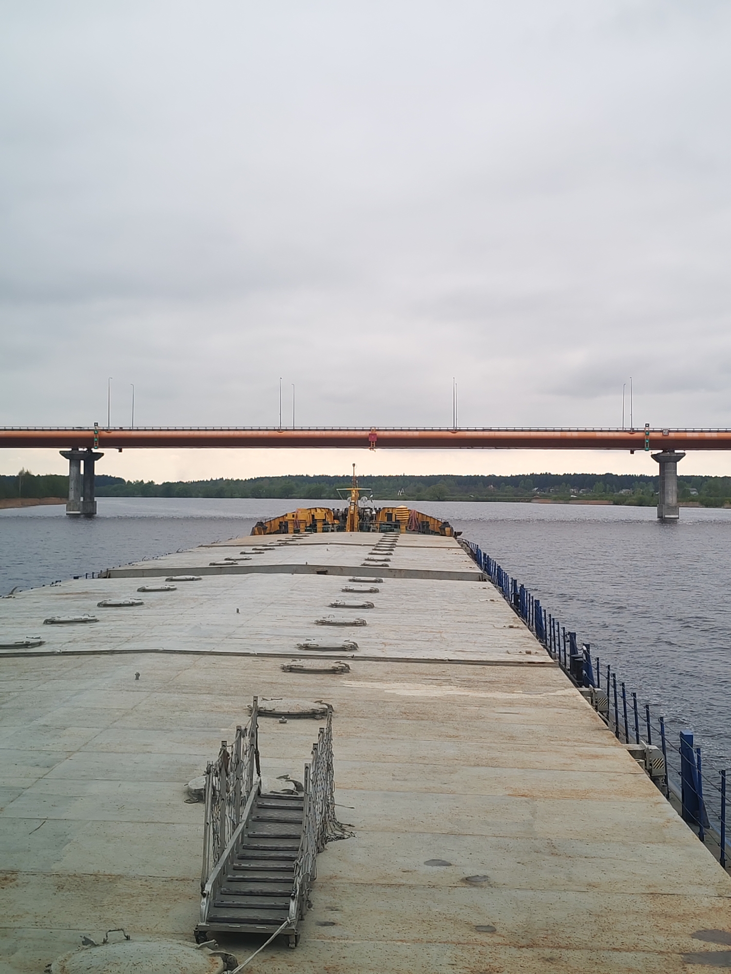 The image depicts a view from a barge on the Volga River in Staroe Semenovskoe, Russia. A concrete deck with multiple access hatches covers the barge. A portable metal staircase is positioned on the deck. Blue railings line one side of the barge. In the background, the Timiryazevsky Bridge spans the river, supported by concrete pillars. A single person in red stands on the bridge. The river flows between the barge and the tree-lined shore. The sky is overcast.