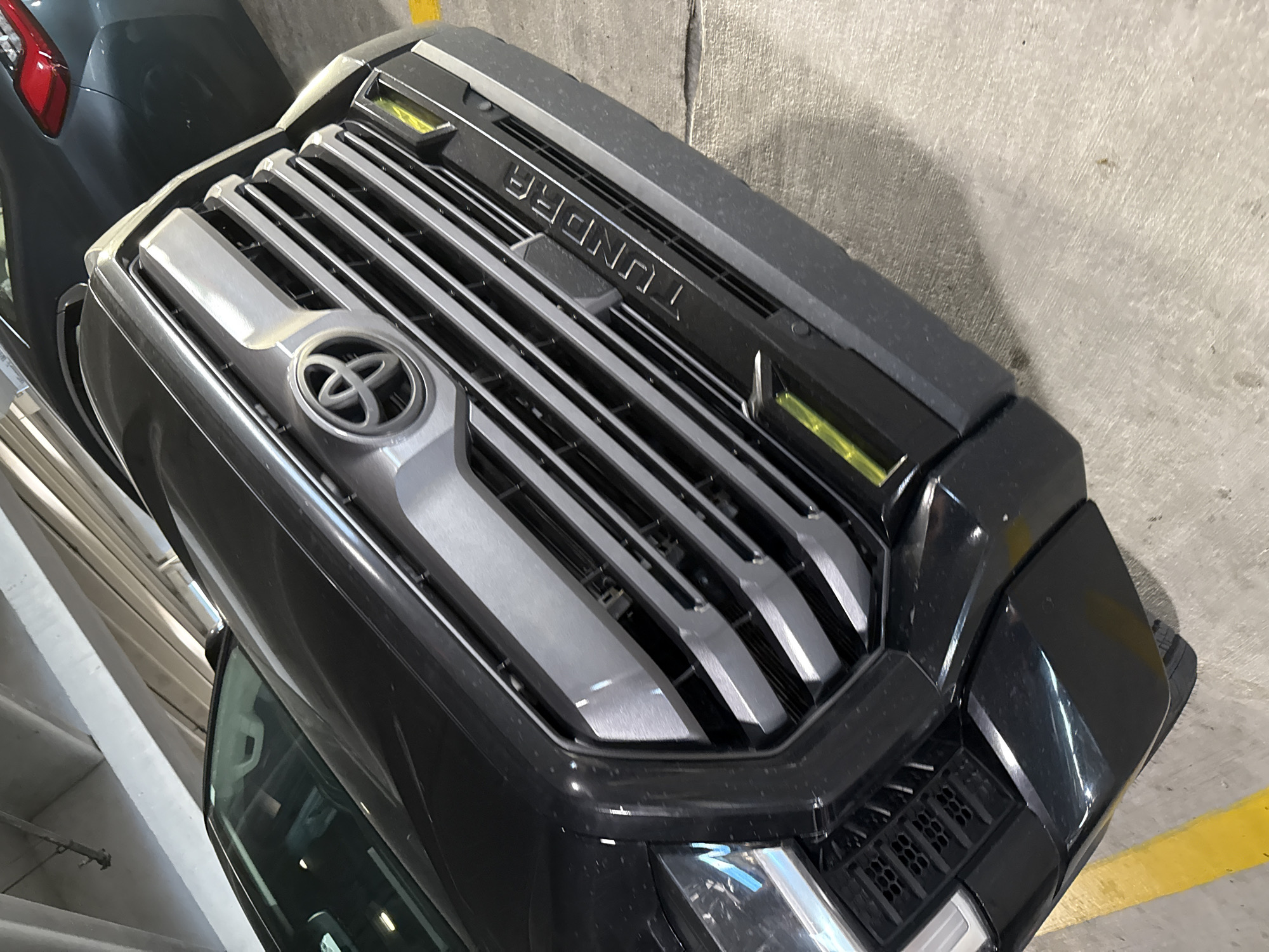 Captured in what appears to be a parking garage in the Hollywood Seminole Reservation, United States of America, the image focuses on the front grill of a black Toyota Tundra. The grill features the Toyota logo, horizontal silver slats, and "TUNDRA" lettering across the top. The vehicle is parked close to a concrete wall, marked with a yellow parking line. A portion of another vehicle is visible on the left side of the frame. The lighting conditions suggest an indoor environment.