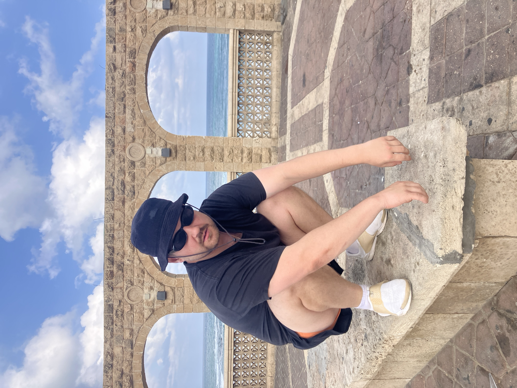 A man is crouching on a stone ledge in front of a stone arched structure and the ocean, in Tel Aviv, Israel. He is wearing a black bucket hat, black sunglasses, a black t-shirt, and orange shorts. He has white sneakers on with white socks. The stone structure has three arches, and behind it, the ocean is visible with a blue sky with white clouds. The ground is paved with bricks in a diamond pattern.