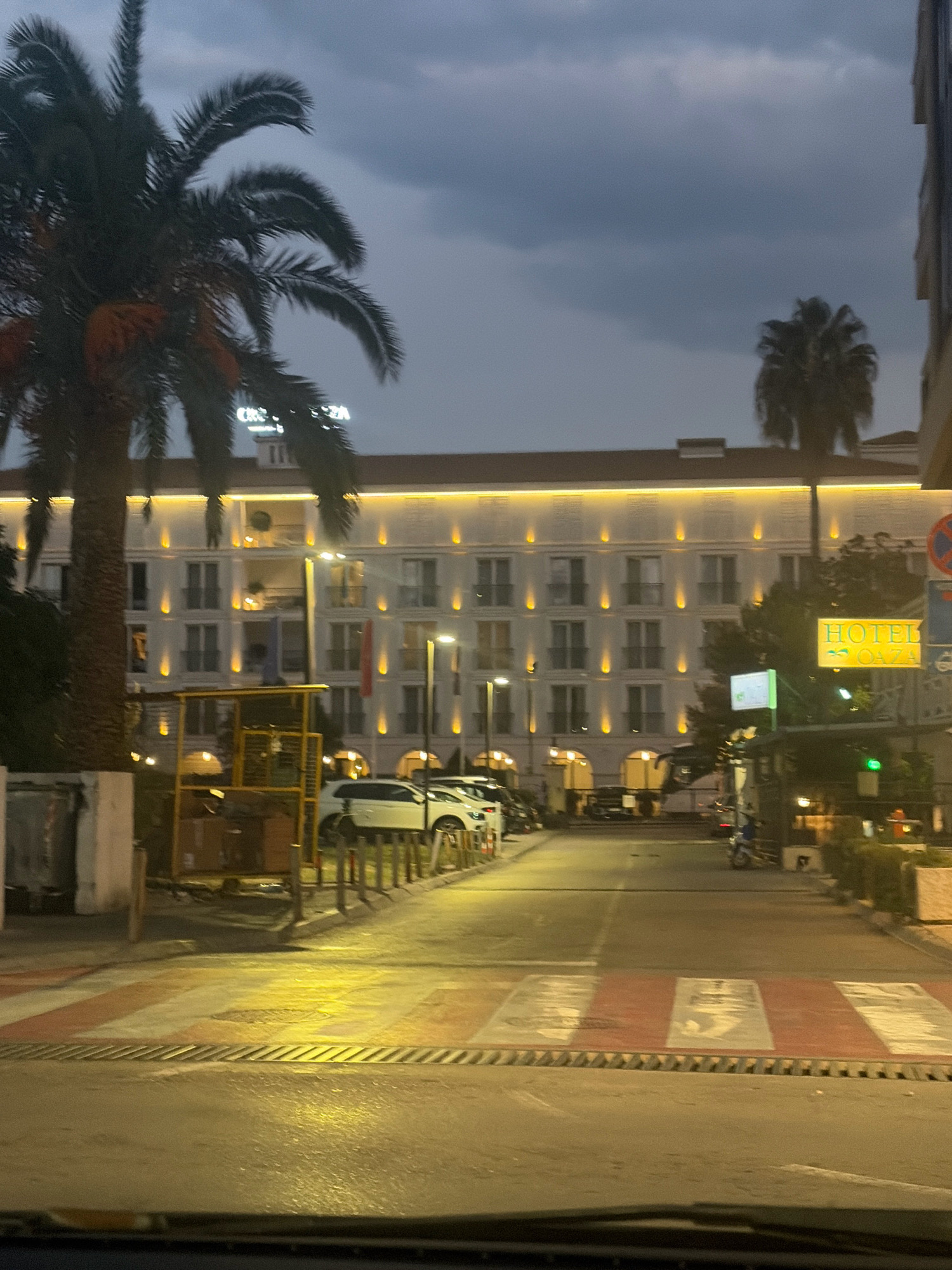 This image depicts the entrance to a hotel in Budva, Montenegro, at dusk. A large palm tree stands to the left, with its fronds partially obscuring the building's facade. The hotel, a multi-story structure with a light-colored exterior, is illuminated by rows of warm yellow lights along its upper edge. Several balconies are visible, some with greenery.
In front of the hotel, a street is lined with parked cars. A yellow sign with the text "HOTEL OAZA" is prominently displayed to the right, along with a smaller green sign. A pedestrian crossing with red and white stripes is in the foreground, indicating a crosswalk. The sky is overcast and dark, suggesting an evening or approaching storm. The overall atmosphere is calm and orderly.